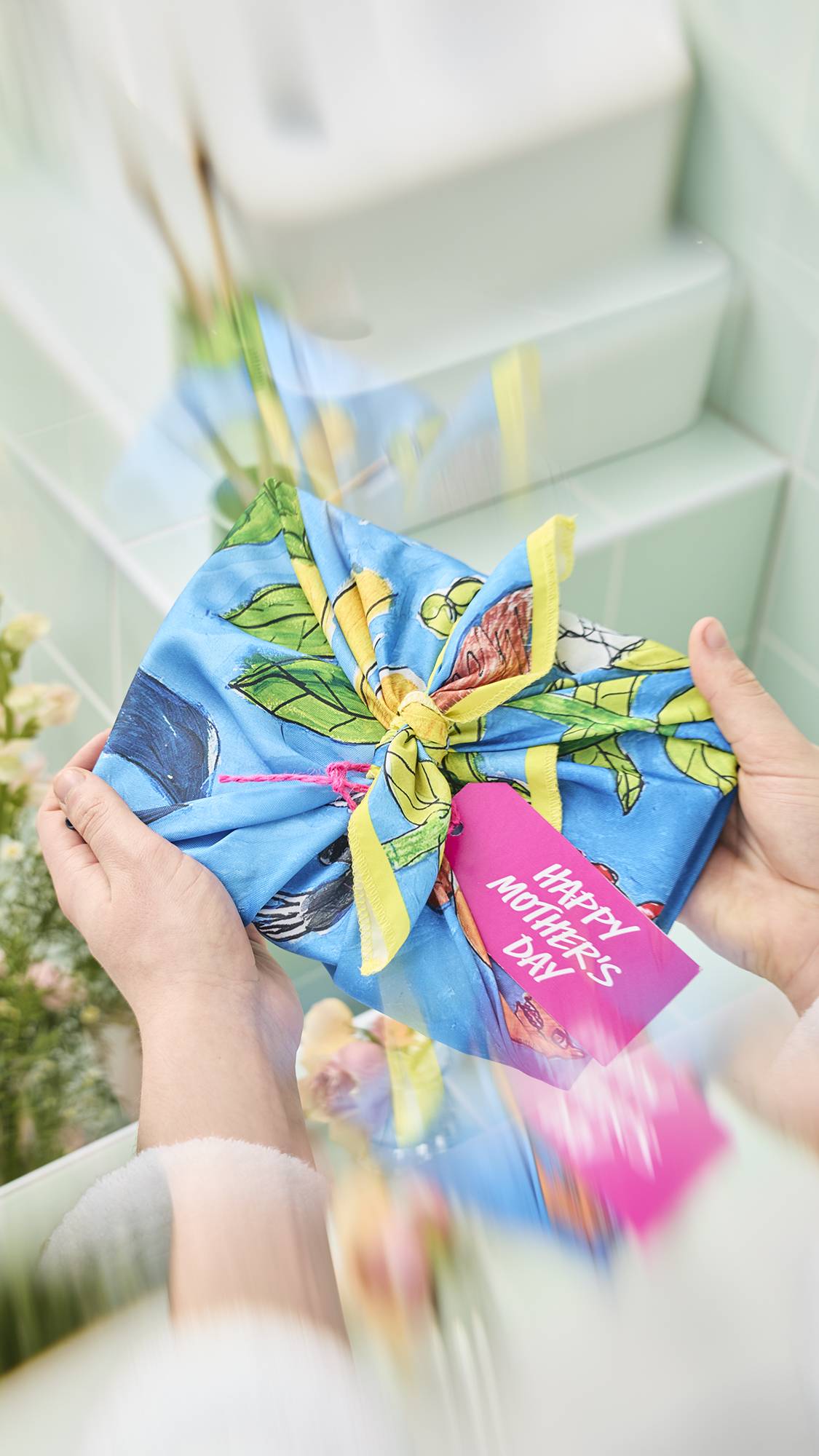 The image shows the arms of a model in a white dressing gown holding a gift wrapped in a "Garden Mural" knot-wrap. The wrap features a whimsical nature design and a pink "Happy Mother's Day" tag is attached to the central knot.