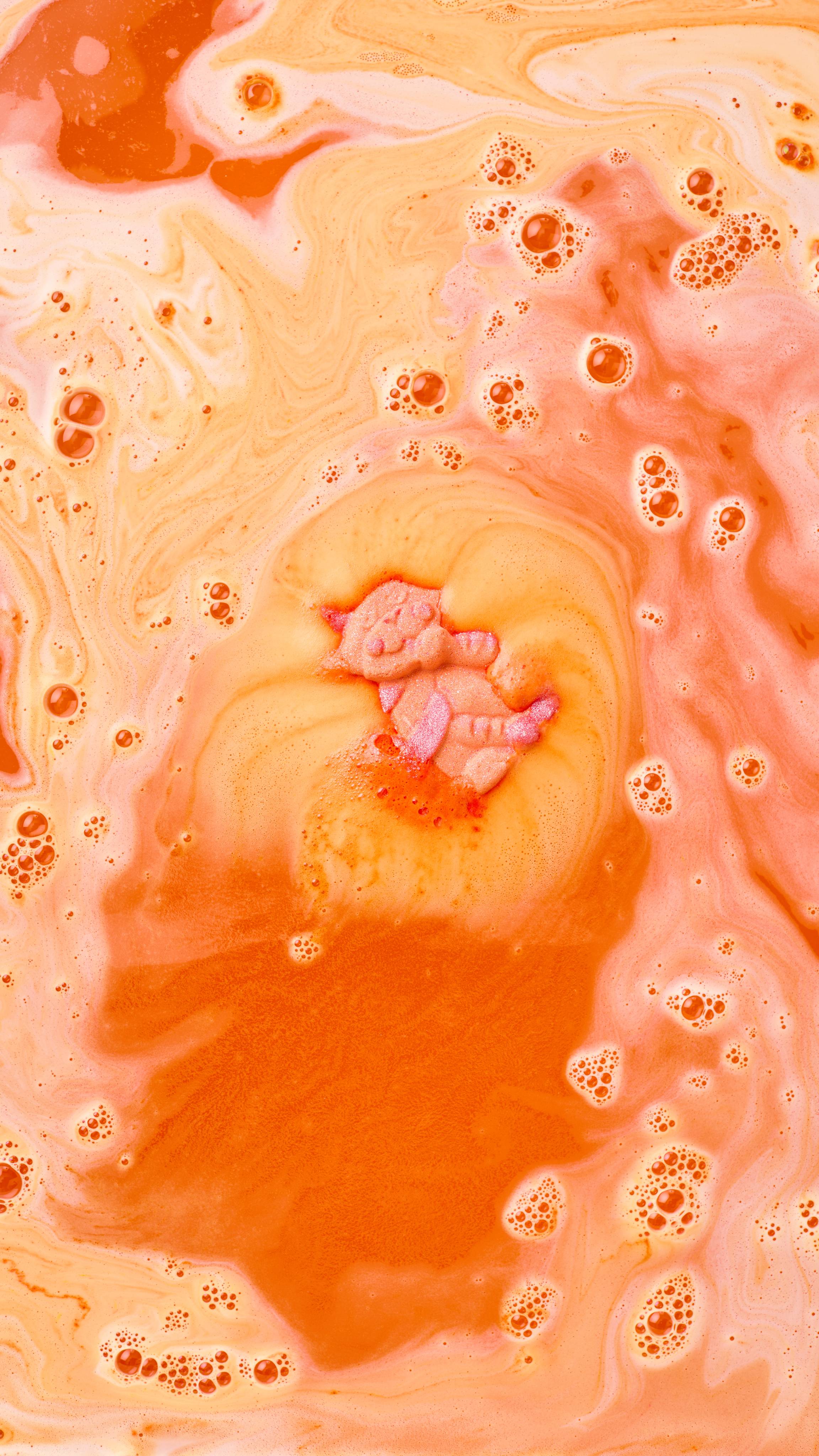 The image shows a vibrant orange 'Ginger Tom' bath bomb, with pink markings, fizzing in swirling yellow and orange bath water. The water forms a colourful spiral around the dissolving cat, blending with thick foam and bubbles.