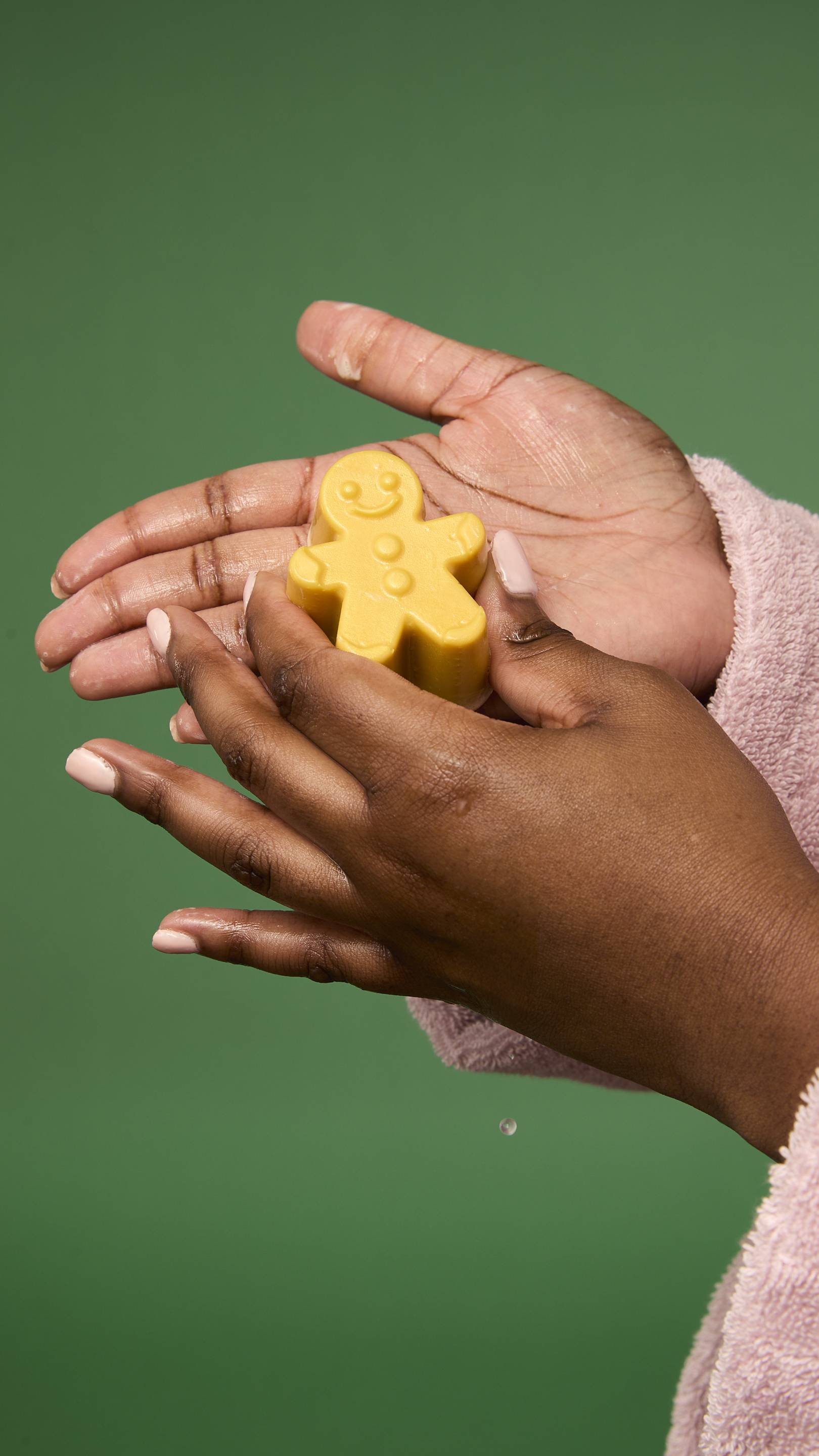 The image shows a dark-skinned model holding a golden-yellow Gingerbread facial gummy wash in their cupped hands. The background is a solid green colour.
