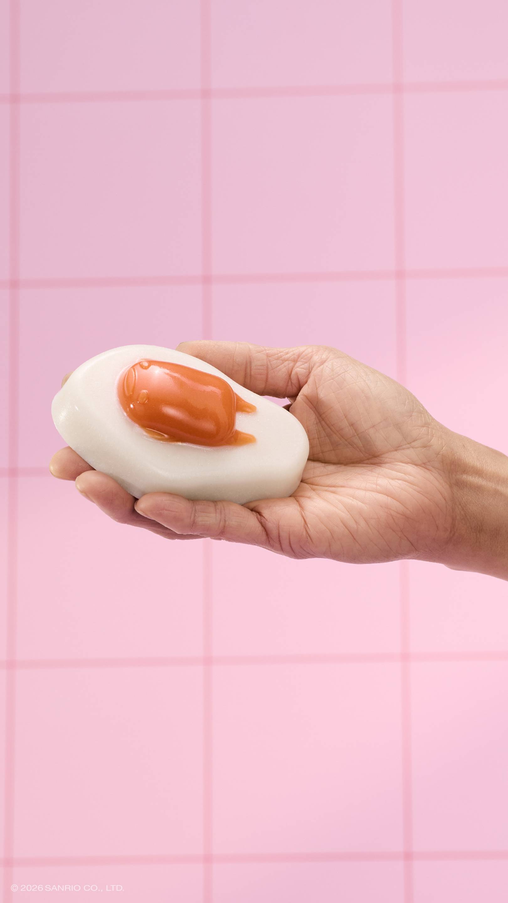 A hand holds a glossy, fried-egg-shaped shower jelly against a background of pink square tiles. The character Gudetama is featured as the orange "yolk" on the white jelly base. 