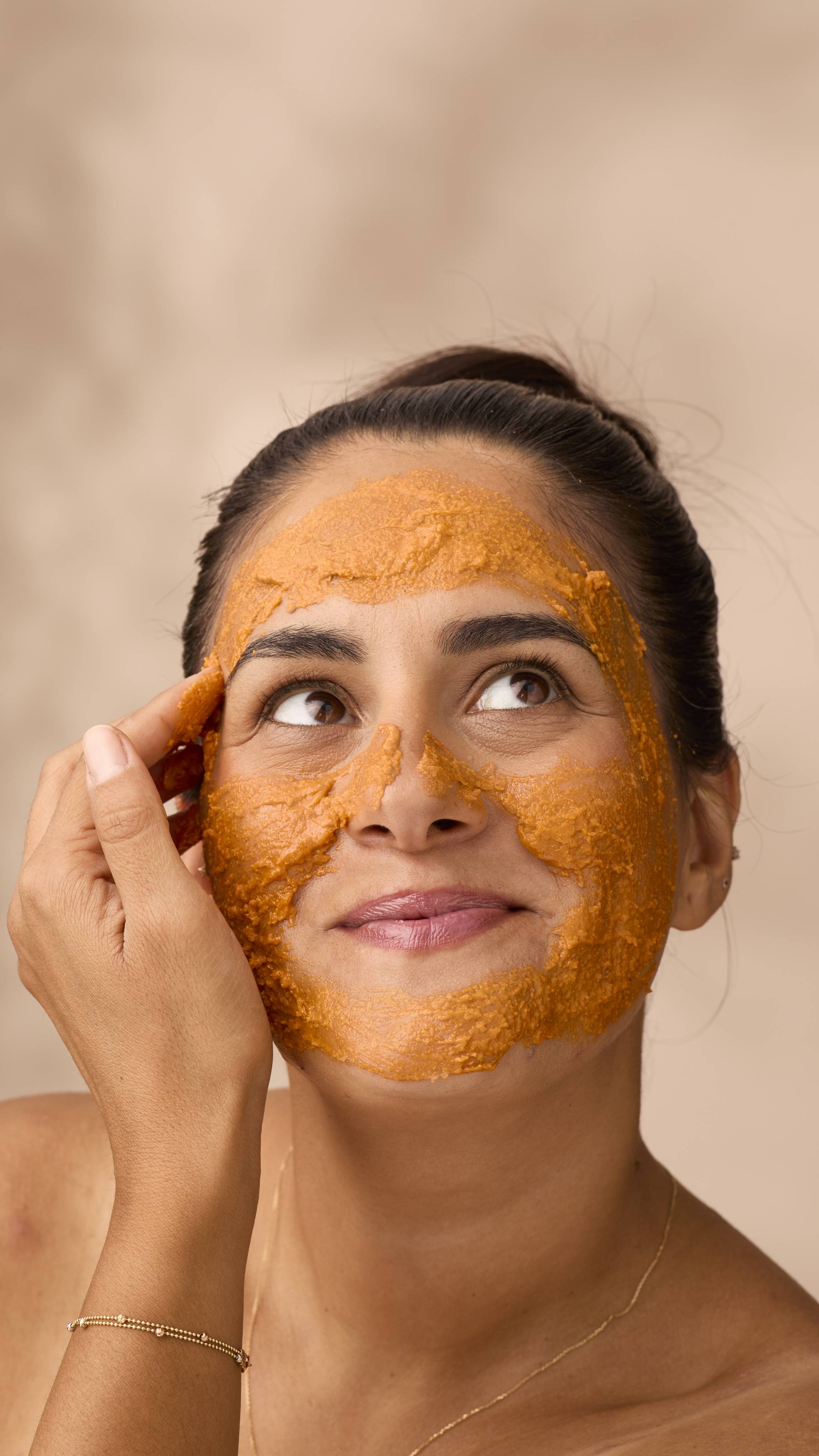A woman with dark hair tied back smiles as she looks to the side, applying a thick layer of bright orange Lush Halloween Queen face mask. The textured paste evenly covers her forehead, nose, cheeks and jawline against a soft warm backdrop