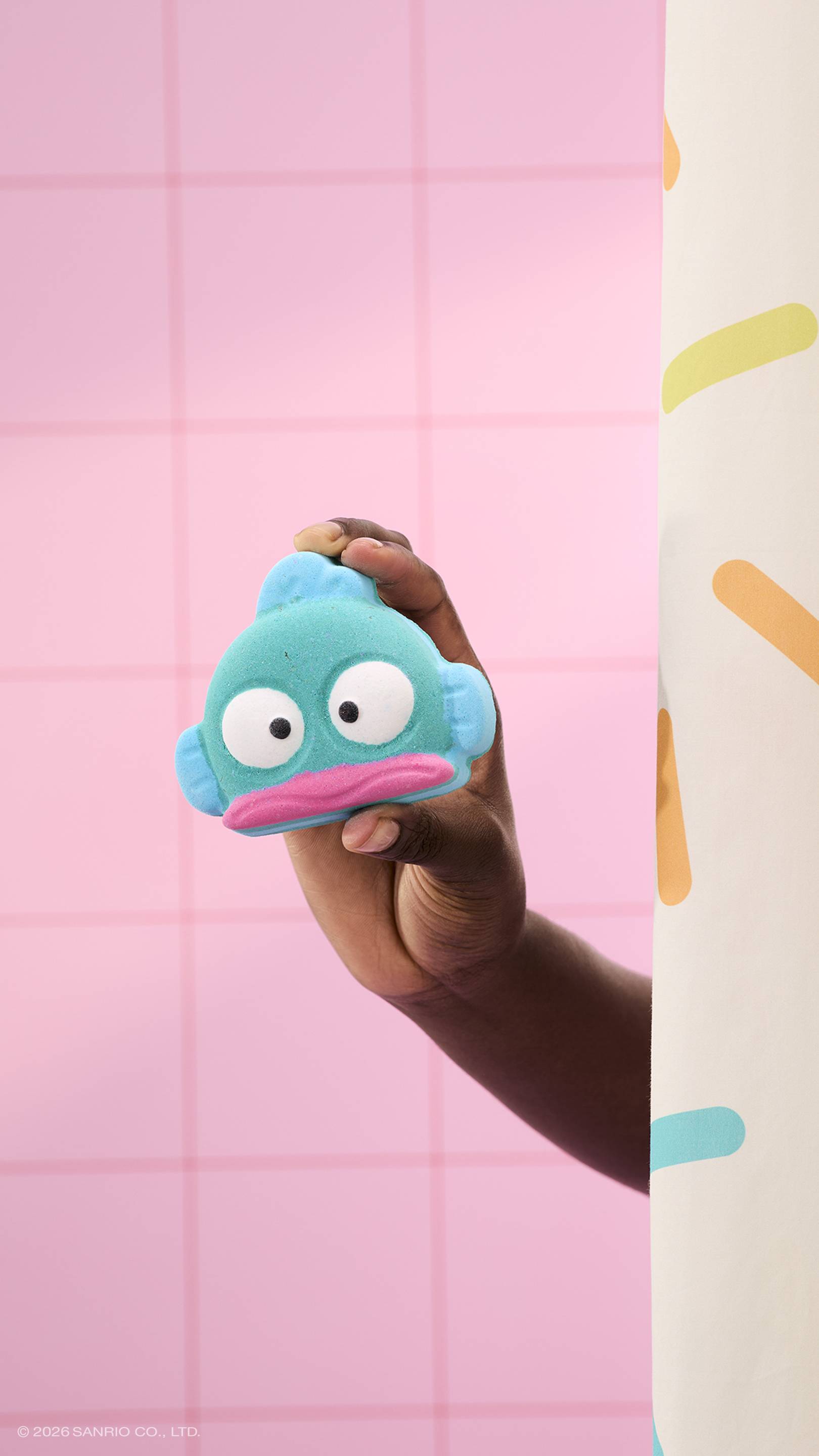 A hand holds a teal, fish-shaped bath bomb with a pink mouth and large white eyes. The person is partially hidden behind a sprinkle-patterned shower curtain, set against a background of pink square tiles.