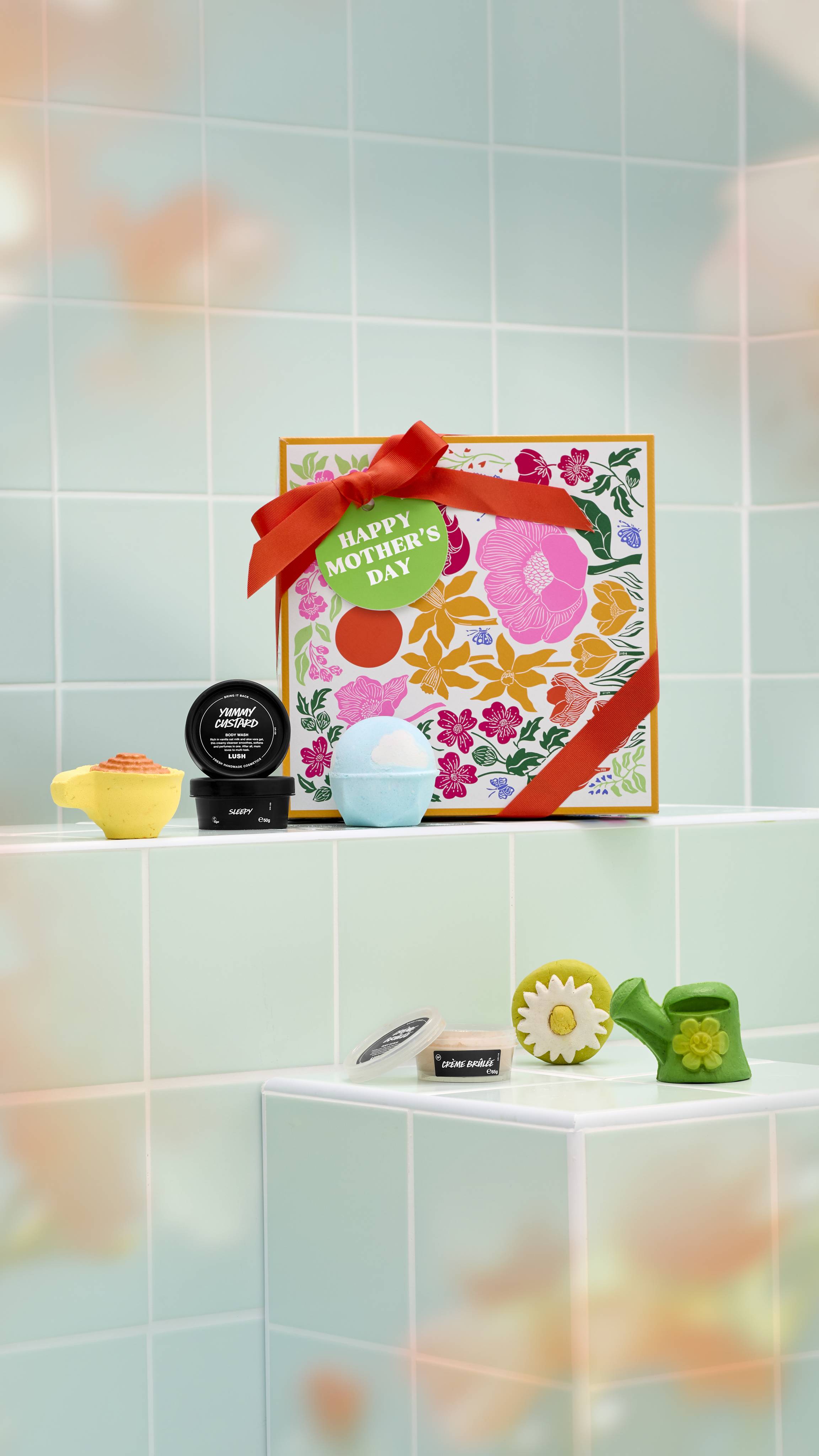 A square Lush "Happy Mother's Day" gift box sits on a pale green tiled ledge. Arranged around it are several bath and body products, including bath bombs, a soap, a bubble bar, and three potted products.