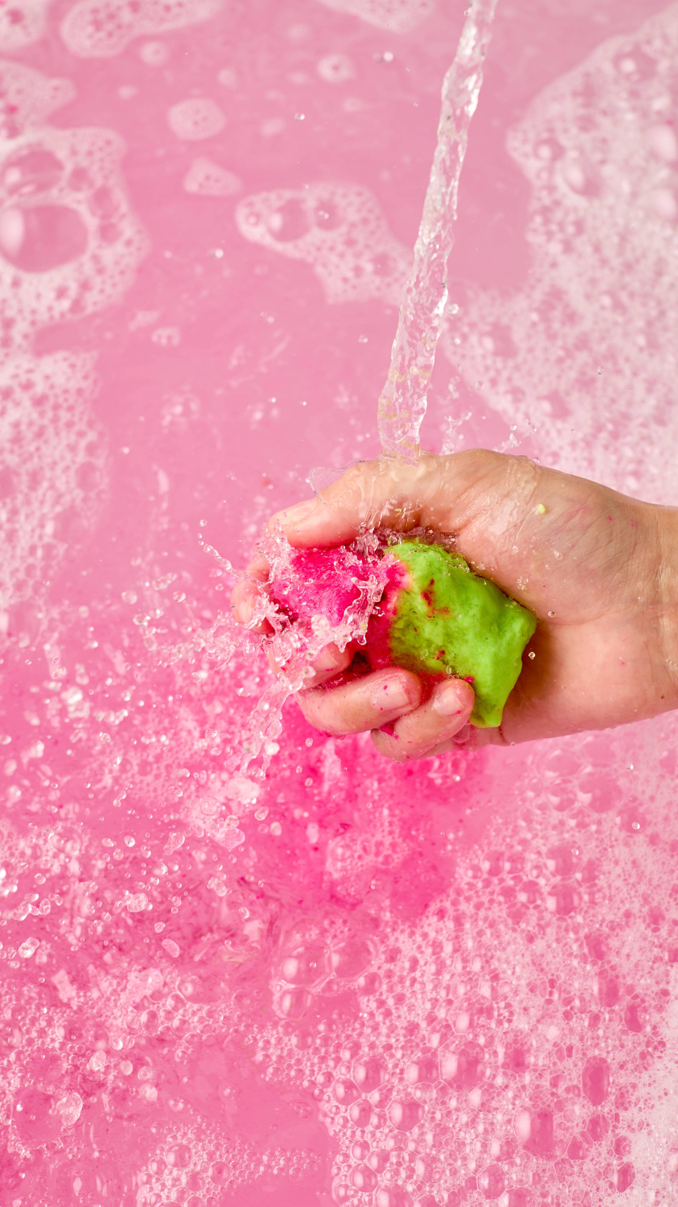 The image shows a light-toned hand vigorously squeezing and crumbling a pink and green 'Invaders from Mars' bubble bar. A strong stream creates vibrant pink bath water with bubbles and thick foam.