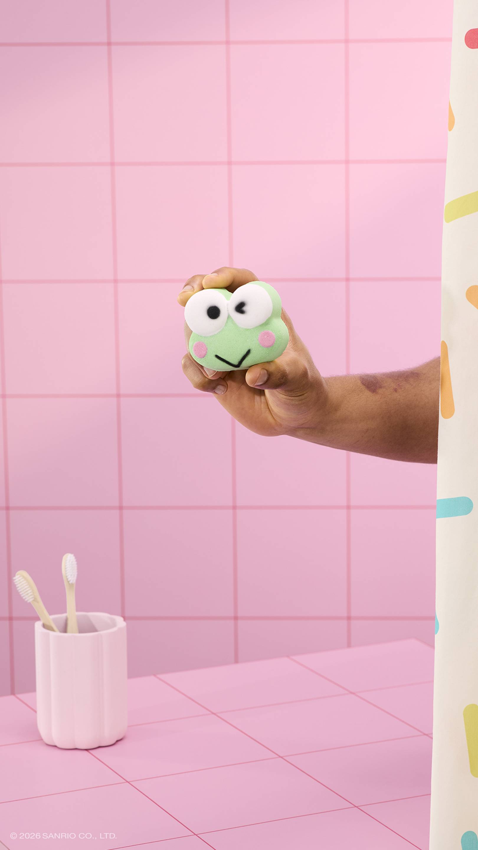A hand holds a green, winking frog-shaped bath bomb in a pink tiled bathroom. A white toothbrush holder with two toothbrushes sits on the pink tiled counter, and a sprinkle-patterned shower curtain is visible on the right. 