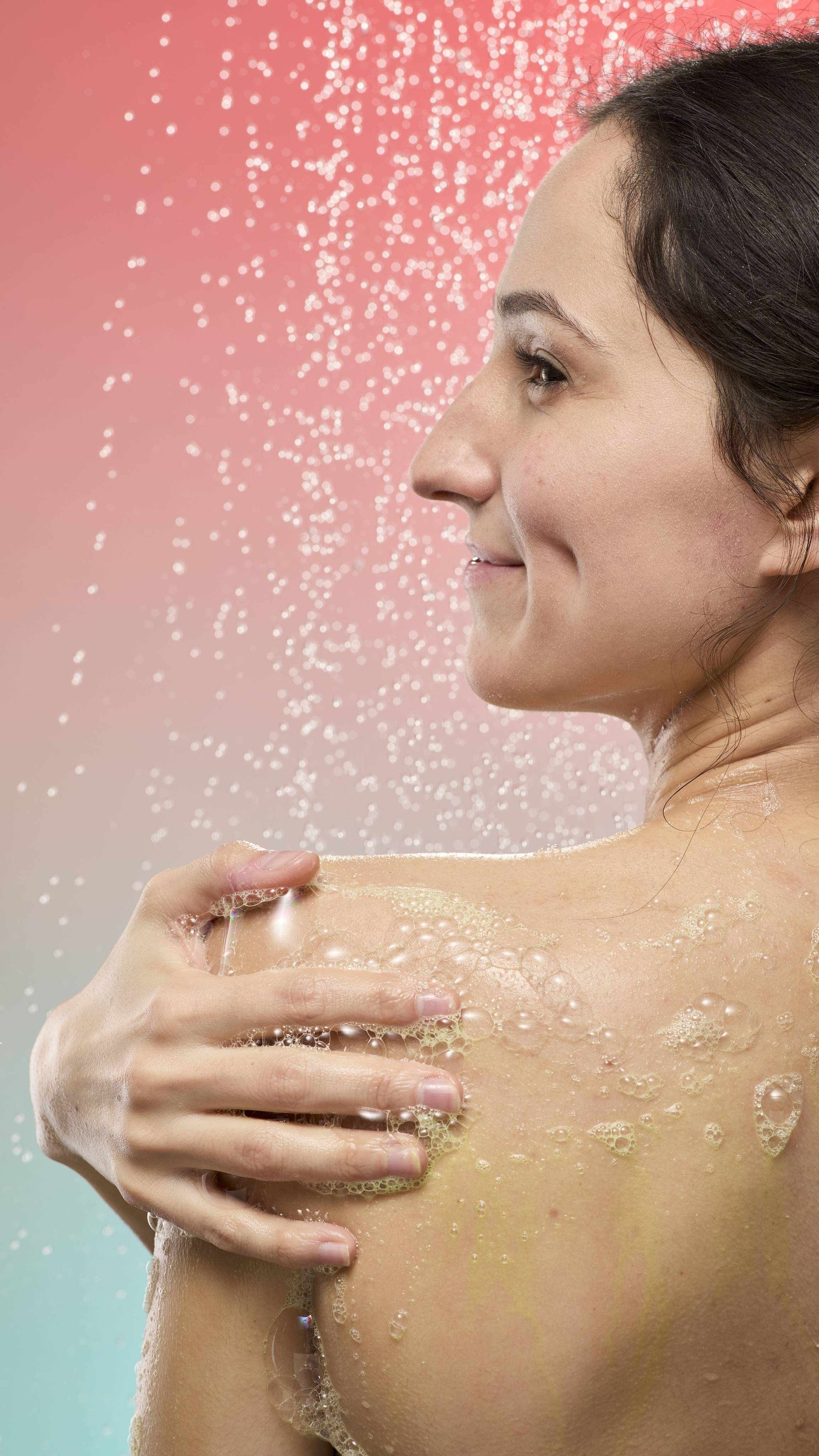 A person smiles in profile while applying foamy, yellowish Lemon Sherbet shower gel to their shoulder. Falling water droplets spray in the background against a soft pink-to-blue gradient.