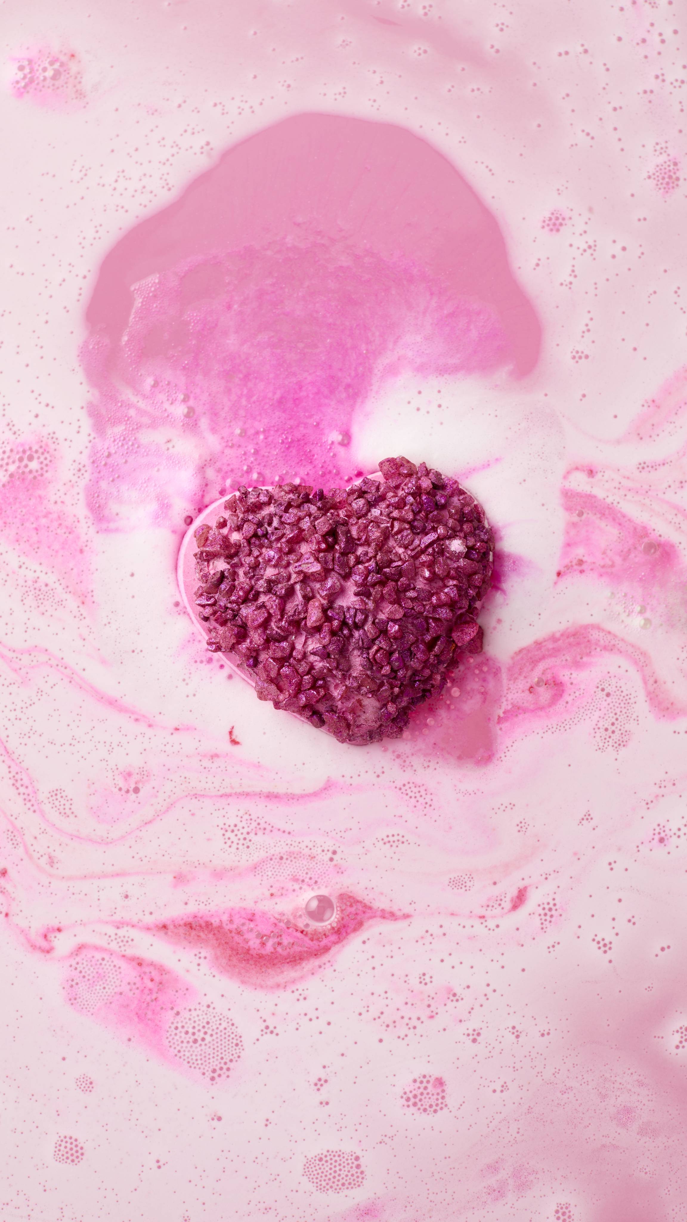 The sparkling magenta heart-shaped Life In Rosy Light bath bomb floats in pale pink water, beginning to dissolve. It leaves a vivid trail of deep pink colour and fine white bubbles as it moves through the swirling bathwater.