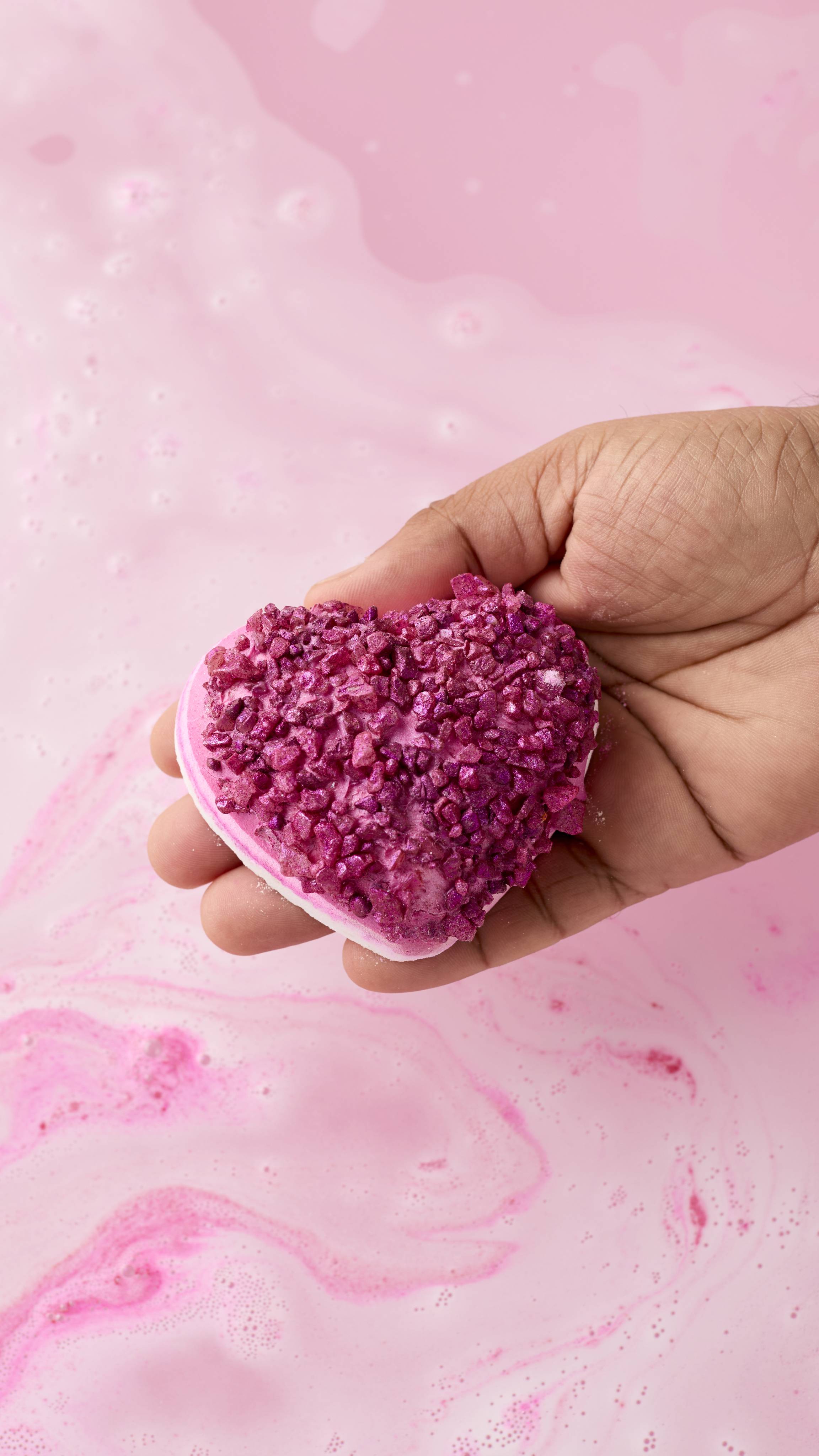 A hand with dark skin holds the pink heart-shaped Life In Rosy Light bath bomb over a tub of pale pink, swirling water. The sparkling magenta crystals on the bomb's surface catch the light against the soft, foamy background.