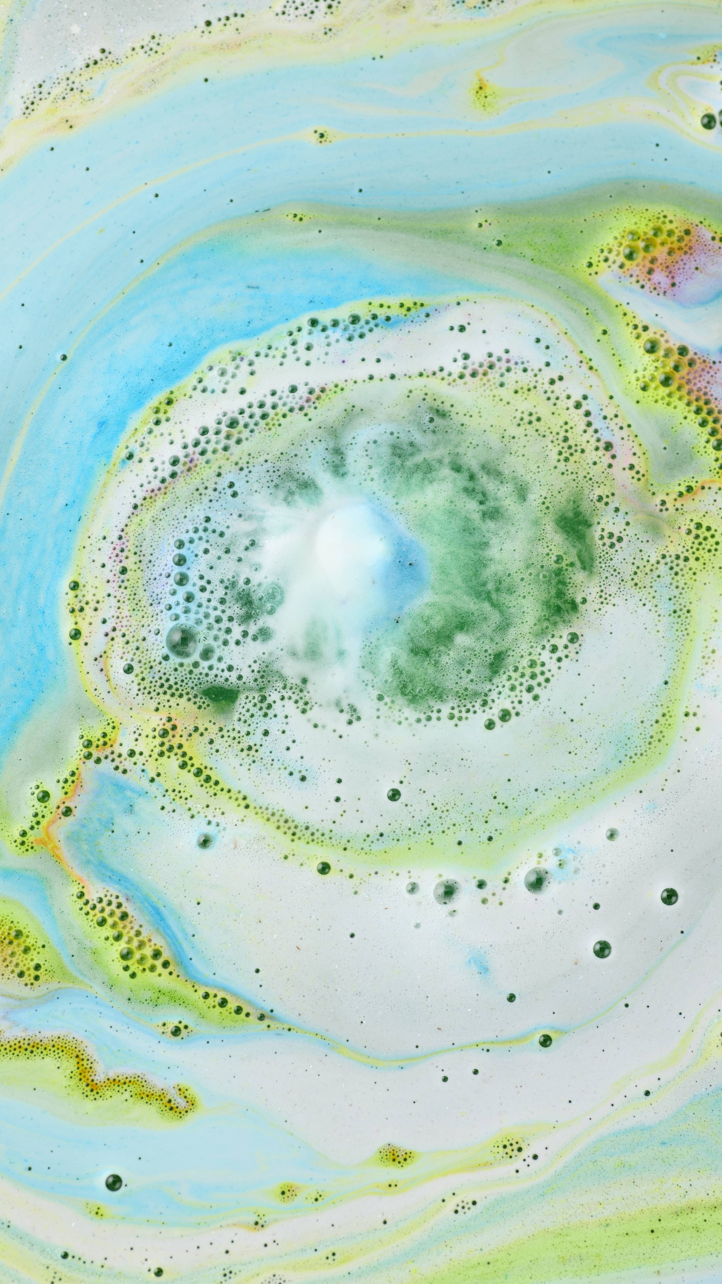 An aerial view of the "Lola's Aurora Borealis" bath bomb dissolving in water, creating a vibrant whirlpool of colour. A central plume of green and white foam radiates outwards into swirling marbled patterns of turquoise, lime green, and pale blue.