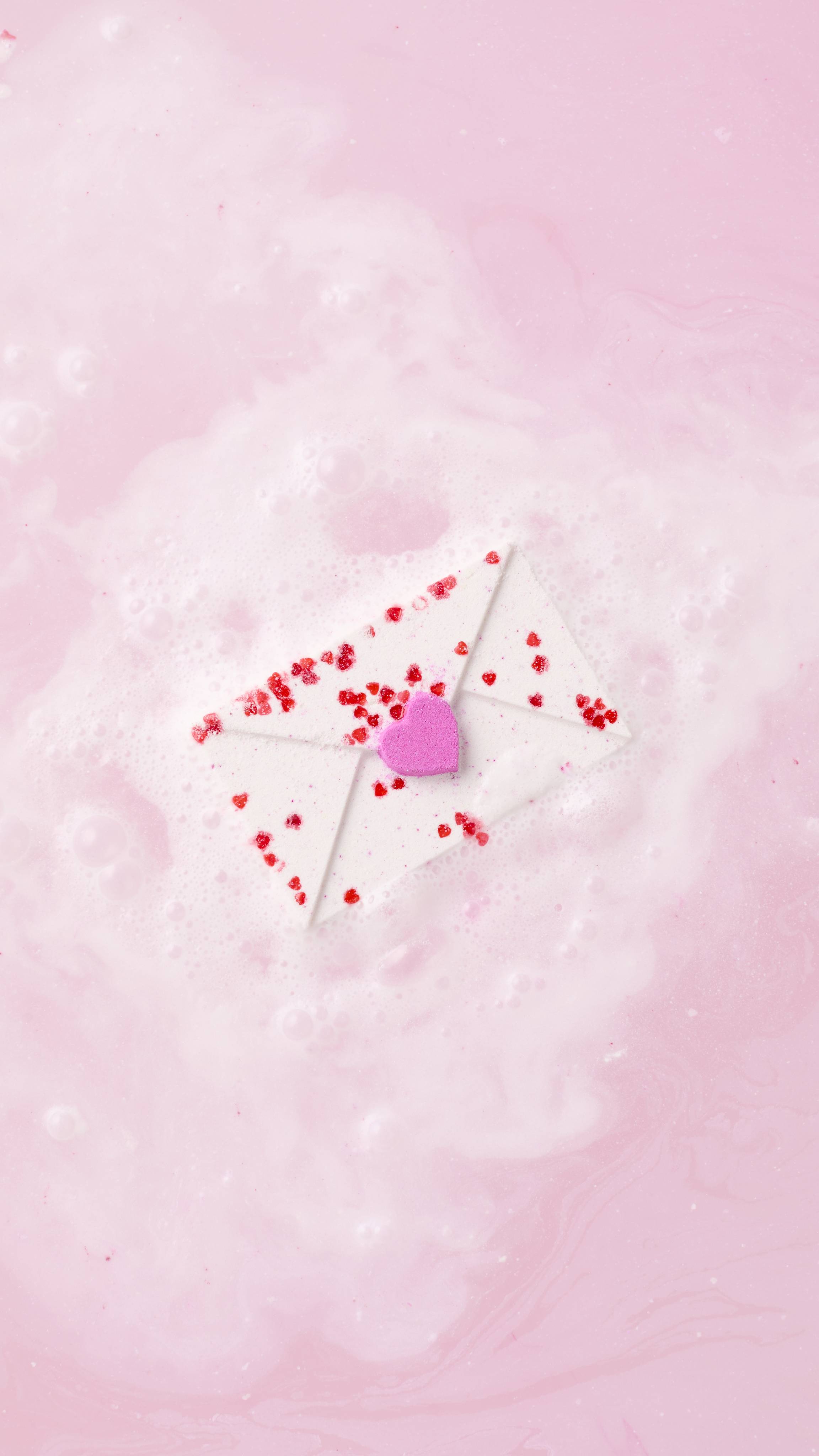 The envelope-shaped Love Letter bath bomb floats in the centre of a bath, surrounded by a cloud of white and pale pink fizz. The water around it is a milky, pastel pink with delicate bubbles on the surface.