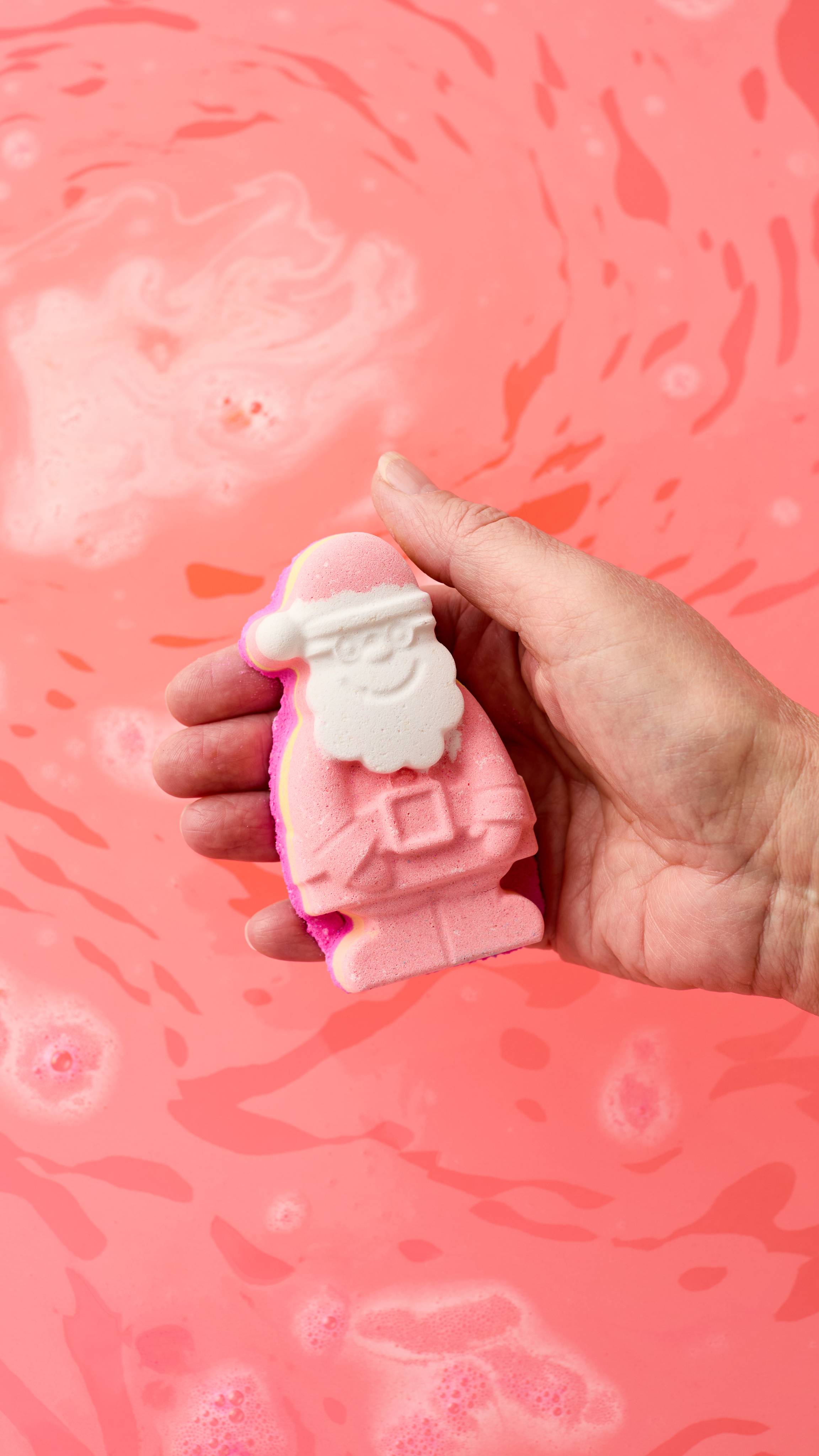 The image shows a hand holding the Santa Claus-shaped, Magical Santa bath bomb. The red suit and hat contrast with the white beard, as the product rests over vibrant pink bath water.