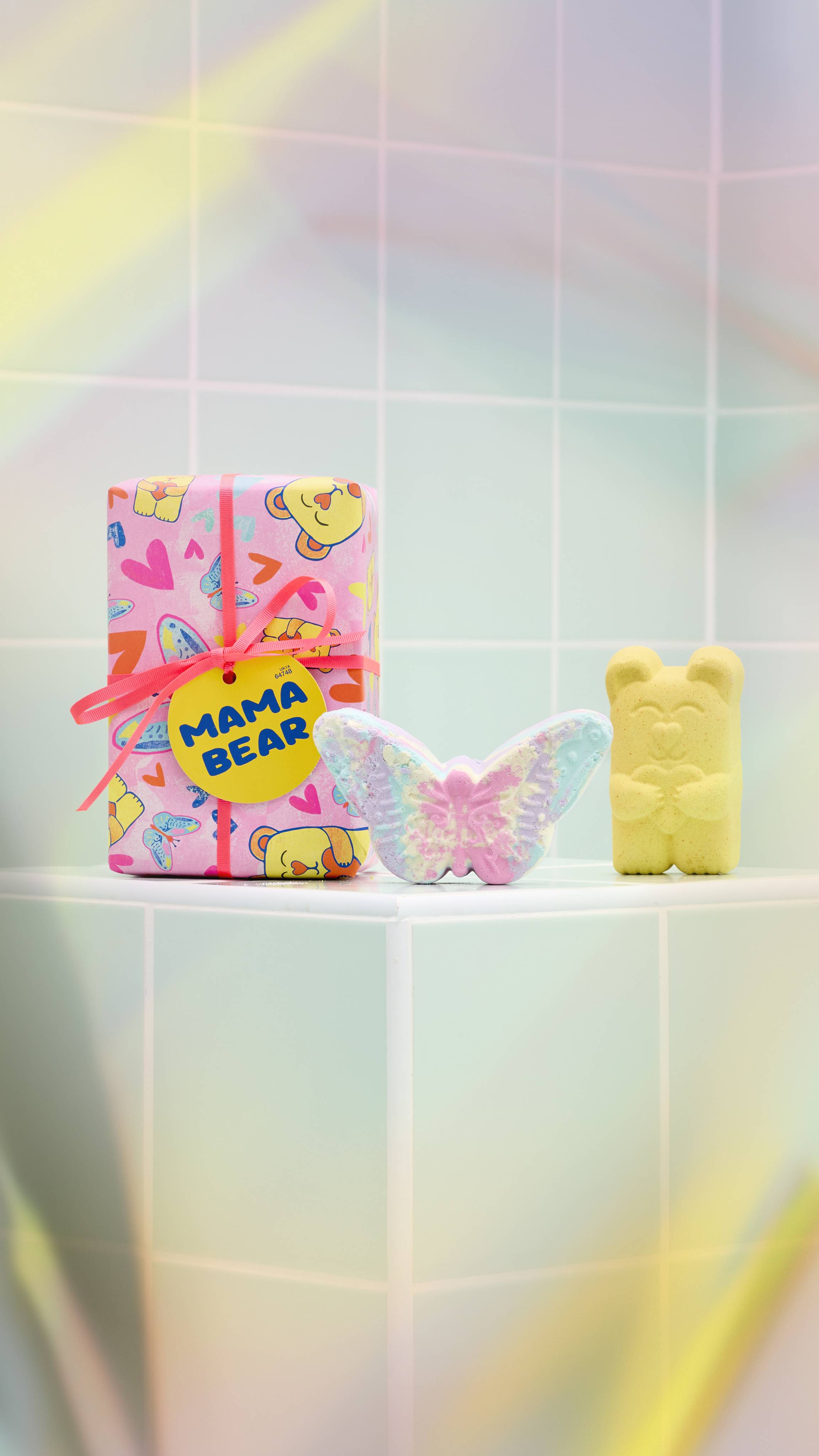 The image shows a brightly wrapped Lush "Mama Bear" gift standing on a pale green tiled ledge. Beside it are a pastel-coloured butterfly bath bomb and a yellow bear-shaped bath bomb. 