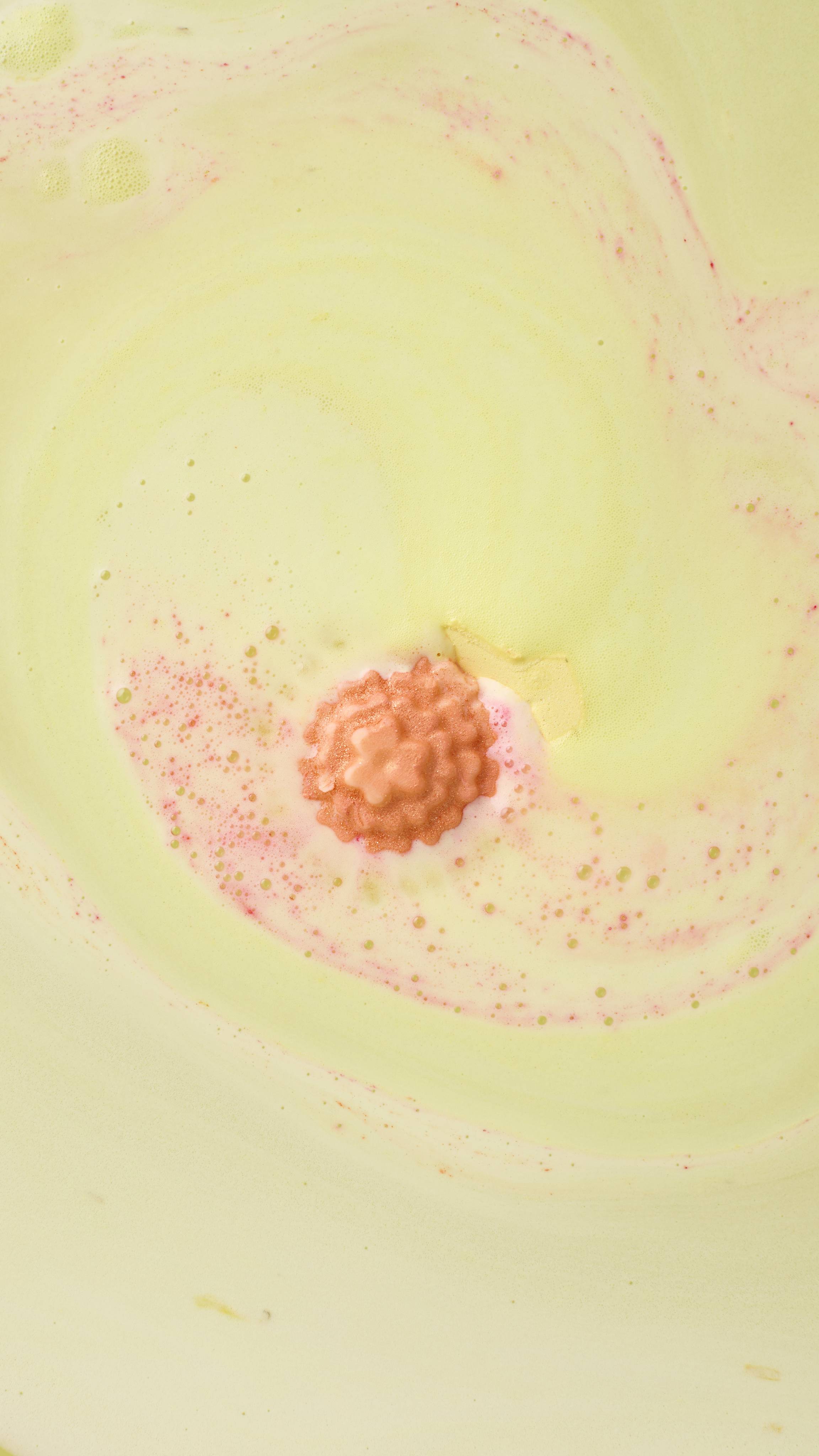 The Marigold Tea bath bomb dissolves, transforming the water into a creamy pale yellow. The textured orange flower center floats on the surface, releasing delicate swirls of pink foam into the milky bath.
