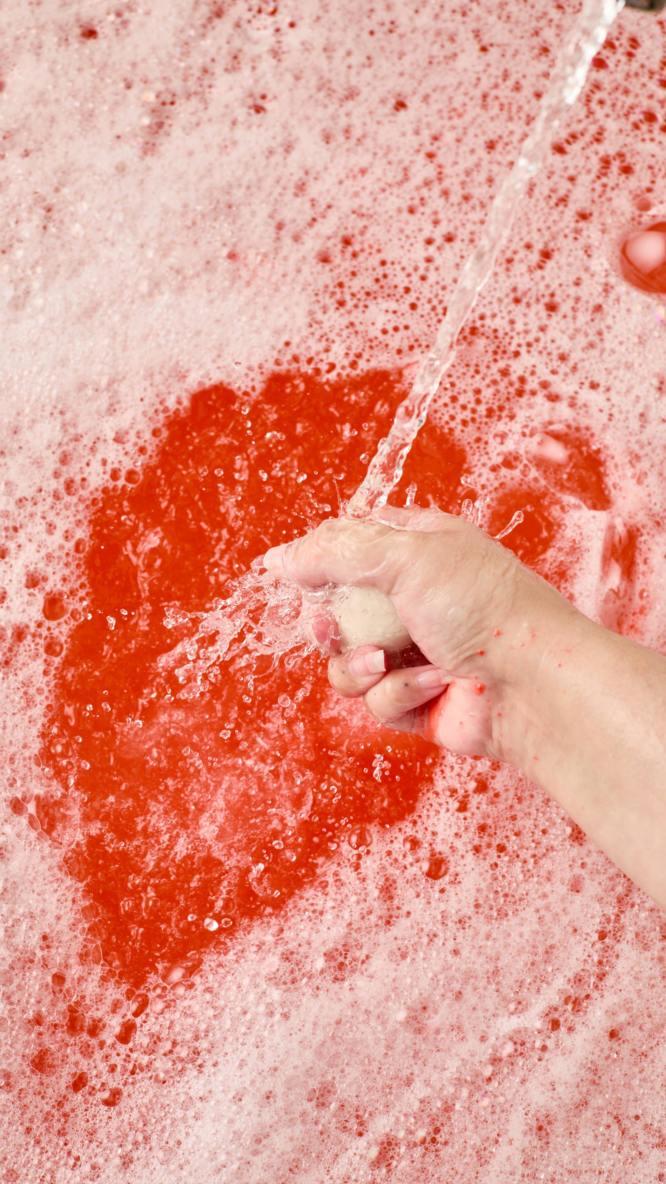 The image shows a light-toned hand vigorously squeezing and crumbling the red, toadstool-shaped 'Midnight Magic' bubble bar under a strong stream of water. Abundant red-pink water is visible under a blanket of bubbles.