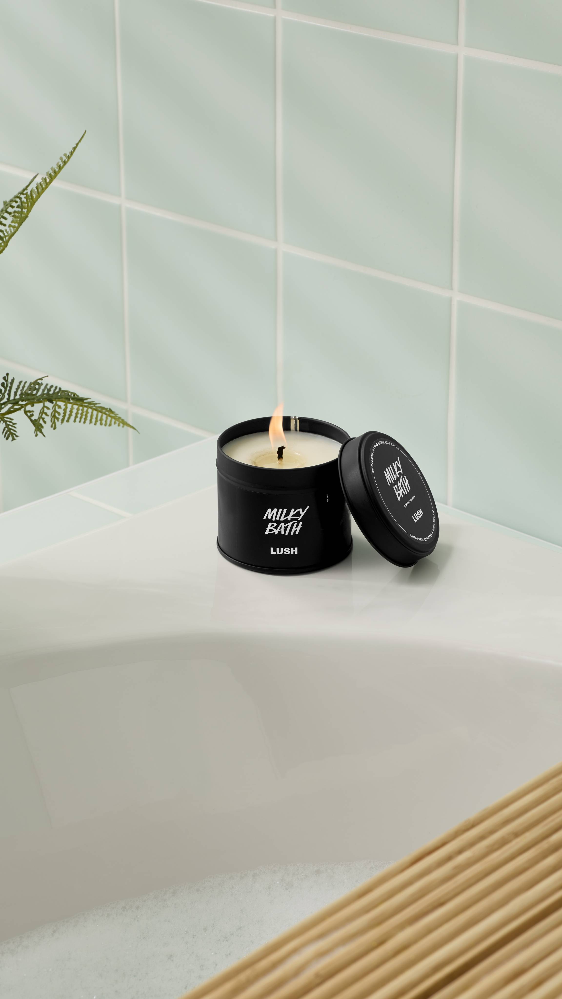 An open, lit Milky Bath candle in a matte black tin rests on the corner of a white bathtub. The lid is propped up against the container. The background features light mint green wall tiles and the leaves of a green fern.