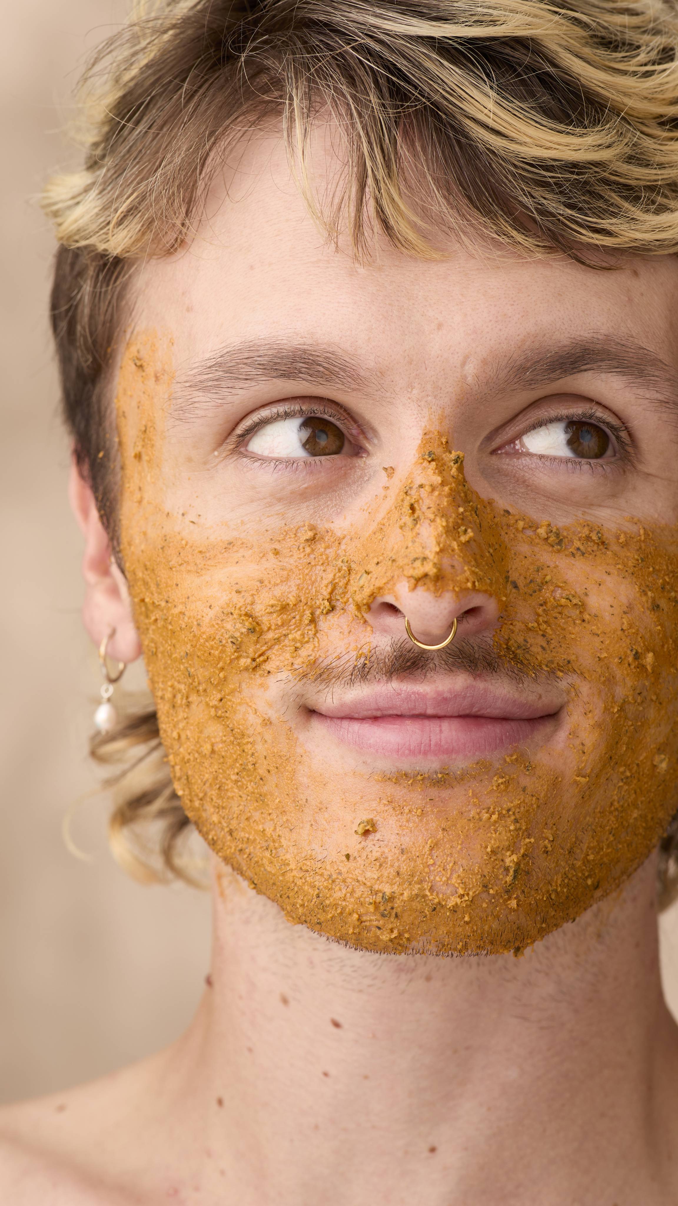 A close-up of a smiling person with blonde-highlighted hair looking to the side, their cheeks, nose and chin covered in a thick, textured layer of orange-brown Lush Mr Pumpkin Face mask. They are wearing a dangling pearl hoop earring.