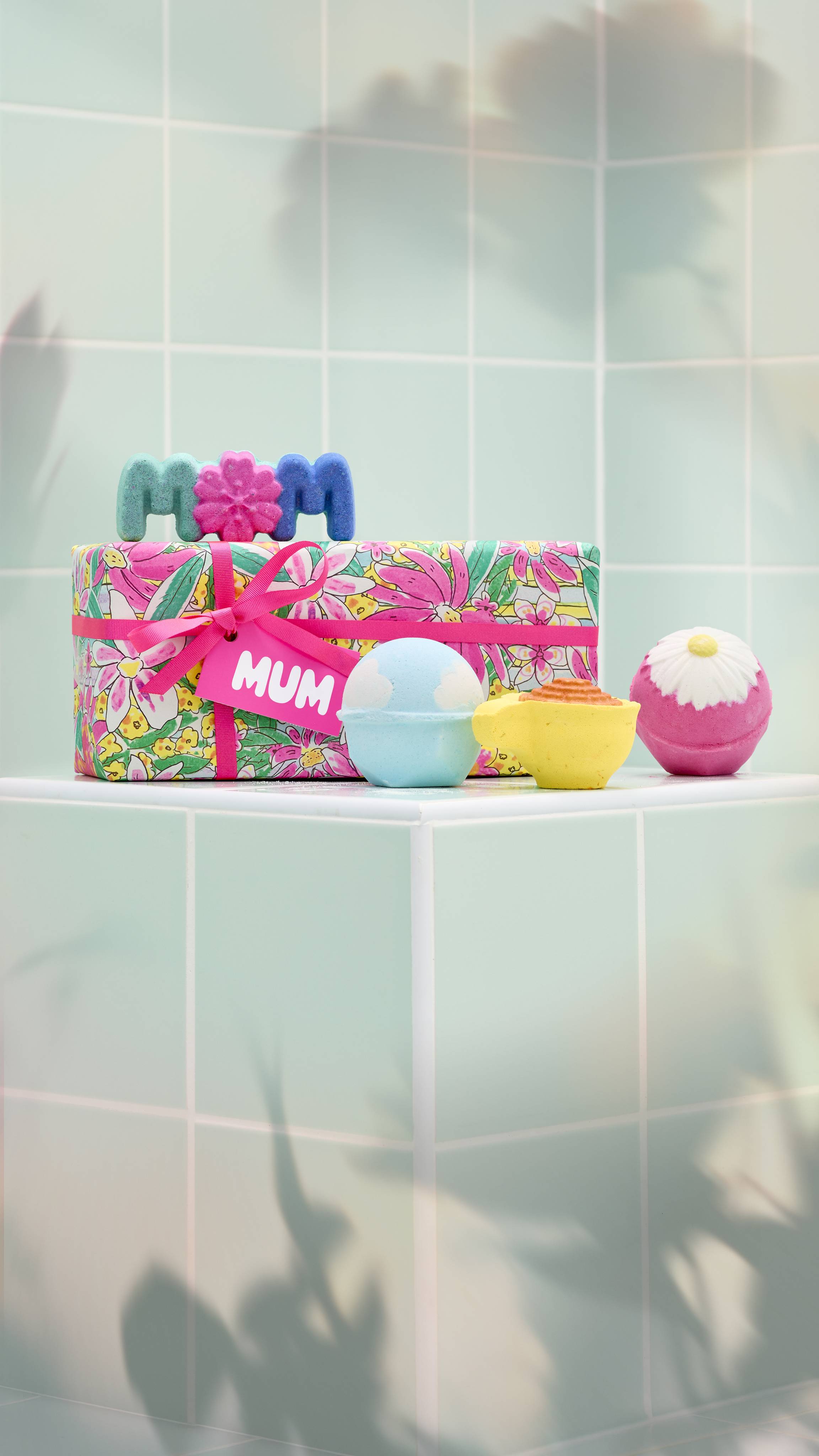 A brightly wrapped Lush "Mum" gift set sits on a pale green tiled ledge. It is accompanied by the four included colourful Mother's Day-themed bath bombs.