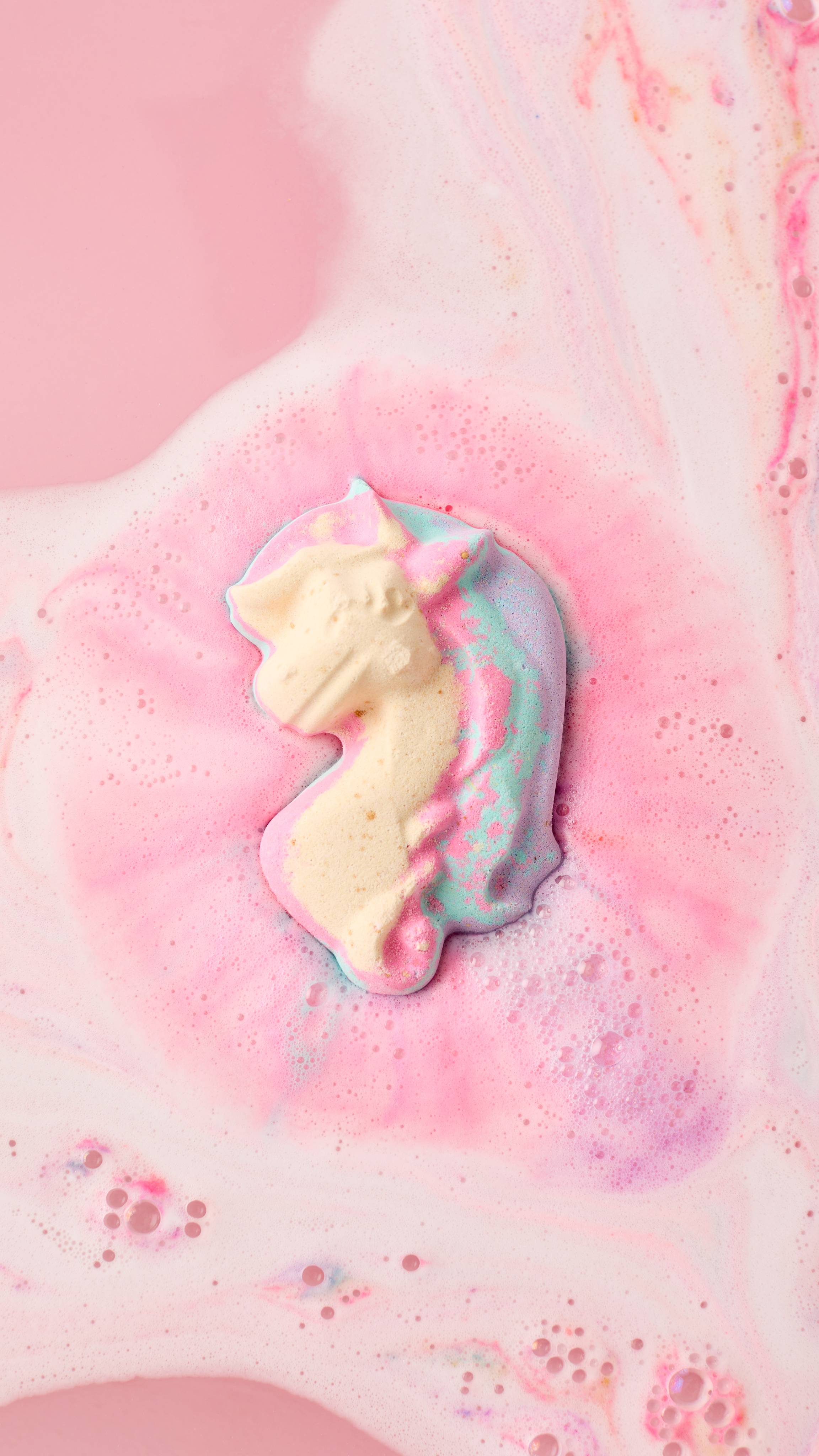 The image shows the My Little Unicorn bath bomb fizzing in a light pink bath. Pastel swirls of pink, purple, and cream radiate outwards from the product, blending with white foam and bubbles.