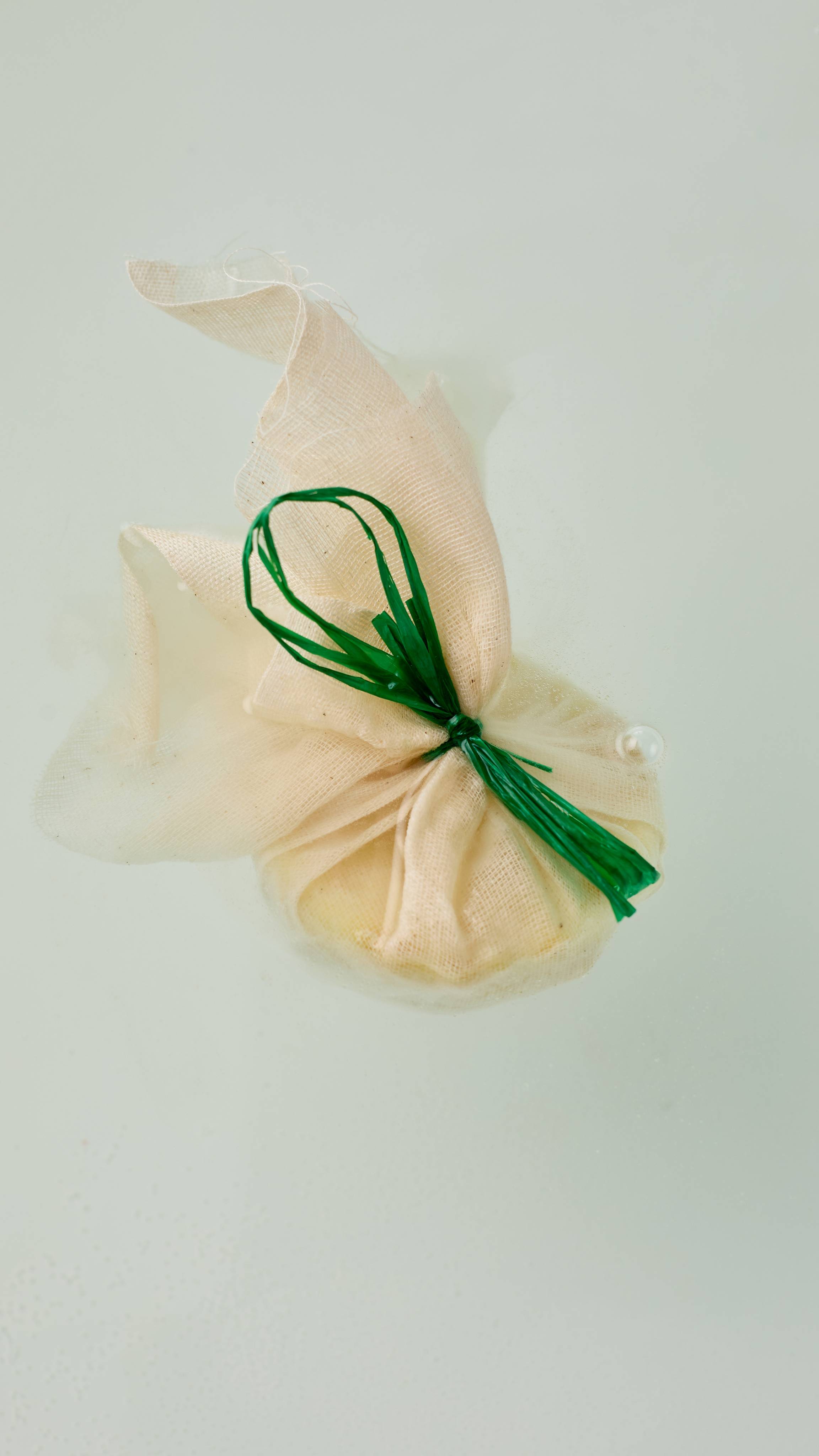 The image shows the Narcotick solid bath oil. A roughly textured muslin fabric pouch, gathered at the middle and tied with a vibrant green string. The top of the fabric flares out above the rounded product, which rests on clear water.