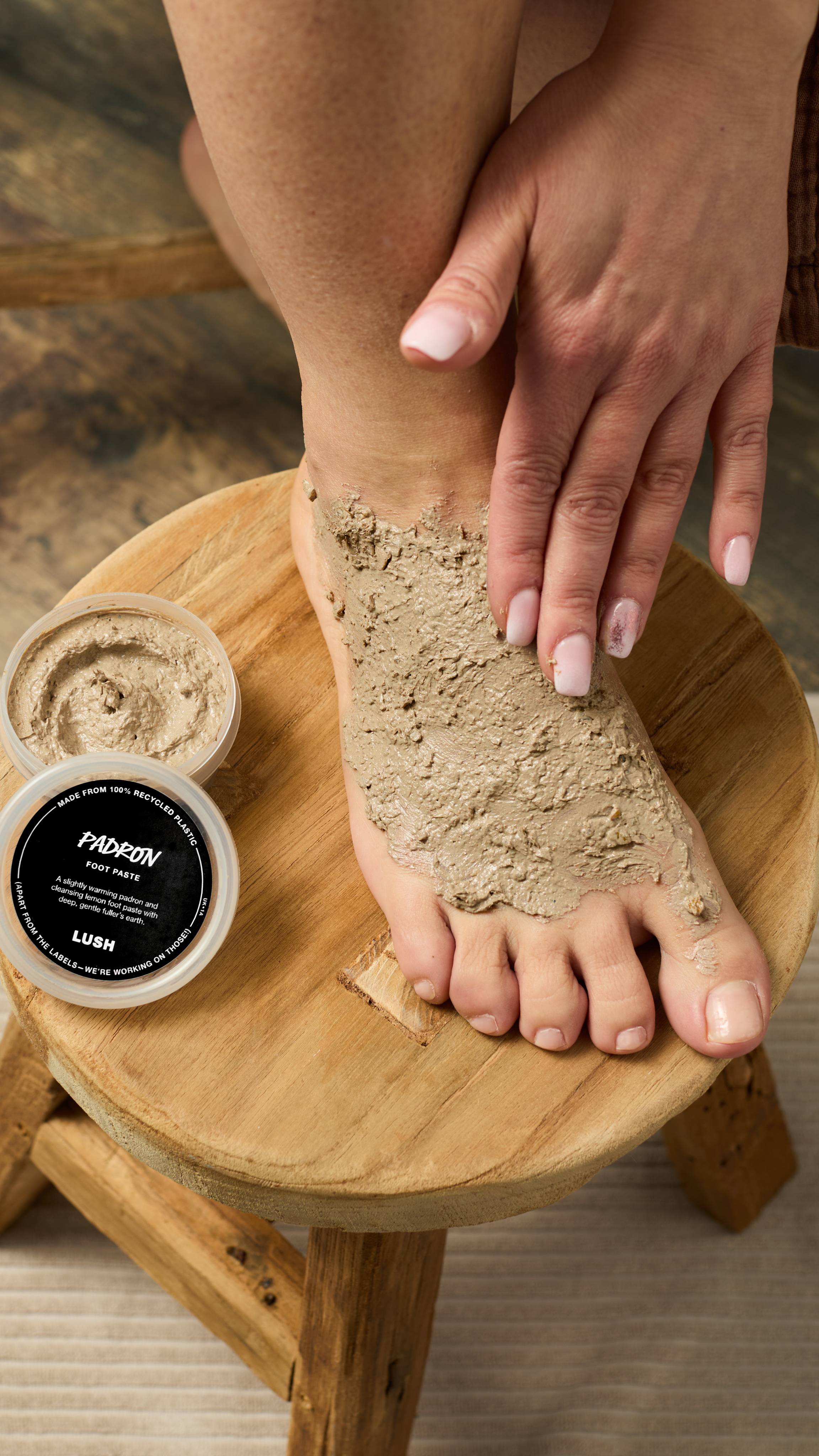 The image shows a hand with light pink nails massaging a thick, dark brown 'PADRON' Foot Mask onto a bare foot, which rests on a small wooden stool. The open mask tub sits nearby, on a woven mat.