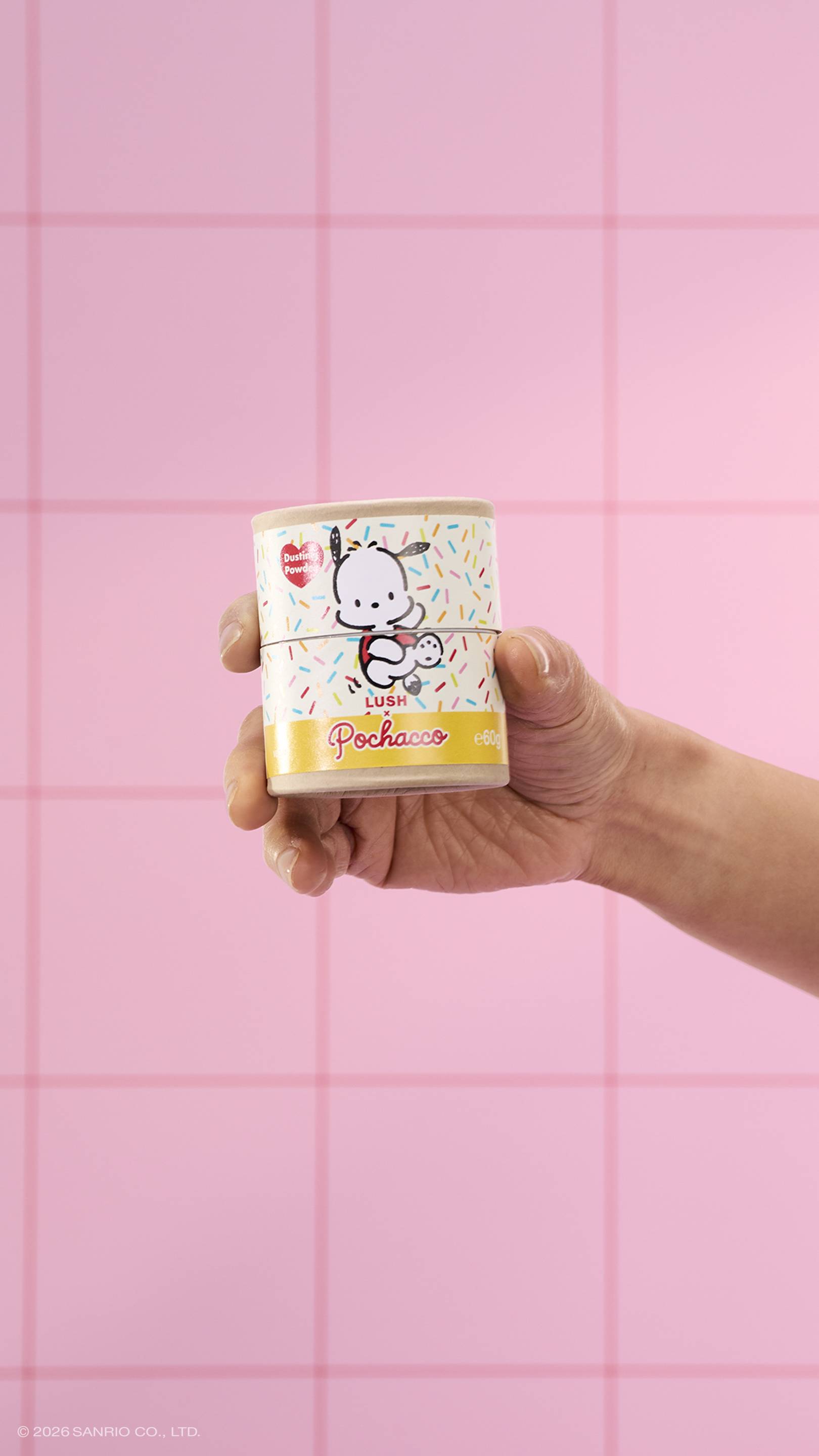 A close-up of a model's hand holding a cardboard tube of Lush x Pochacco dusting powder, featuring a sprinkle pattern and a cartoon dog. The background consists of pink square tiles.
