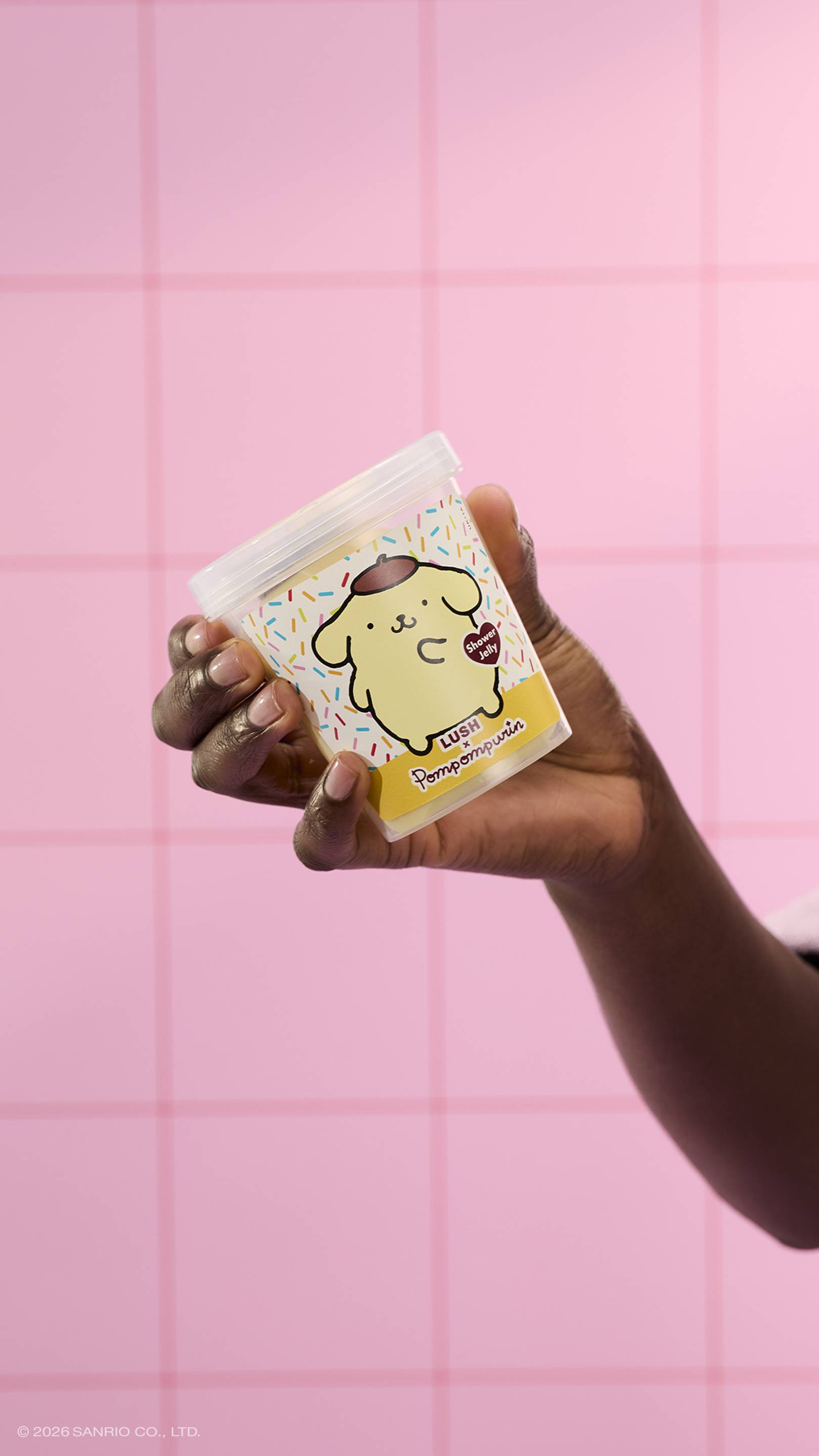 A hand holds a clear plastic tub of Lush x Pompompurin shower jelly, featuring a yellow dog character on the label. The background consists of pink square tiles. 