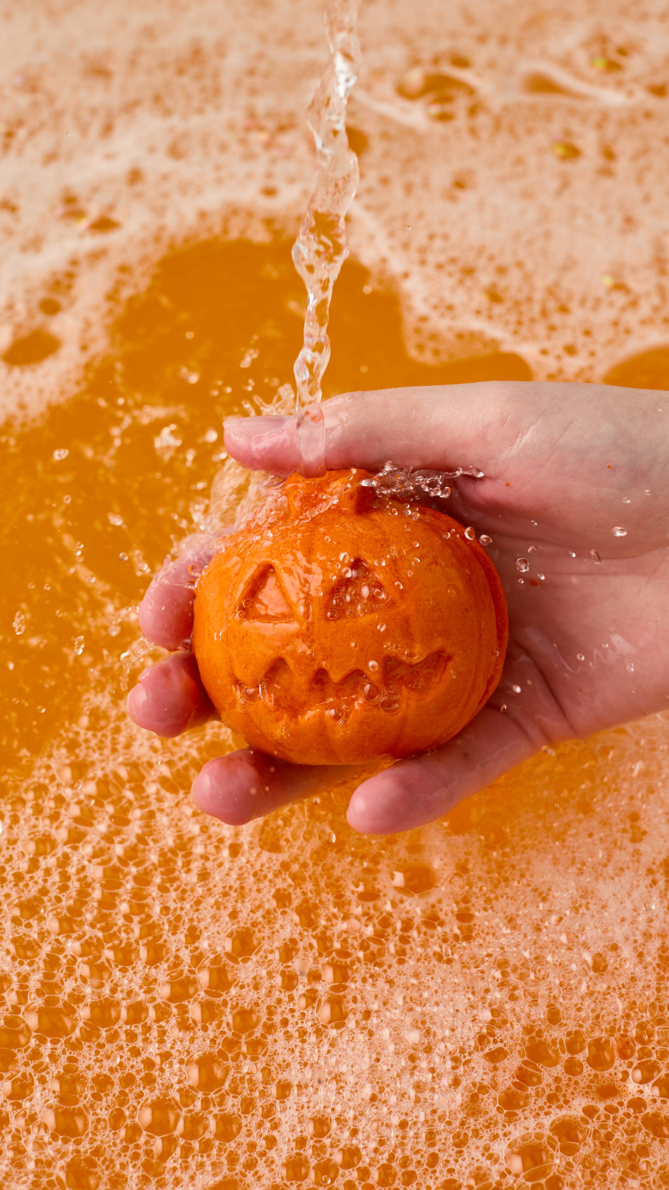 The image shows a light-toned hand holding an orange 'Pumpkin Crumble' bubble bar with carved jack-o'-lantern facial features. Water pours onto the bar, generating abundant bubbles and vibrant orange bathwater.