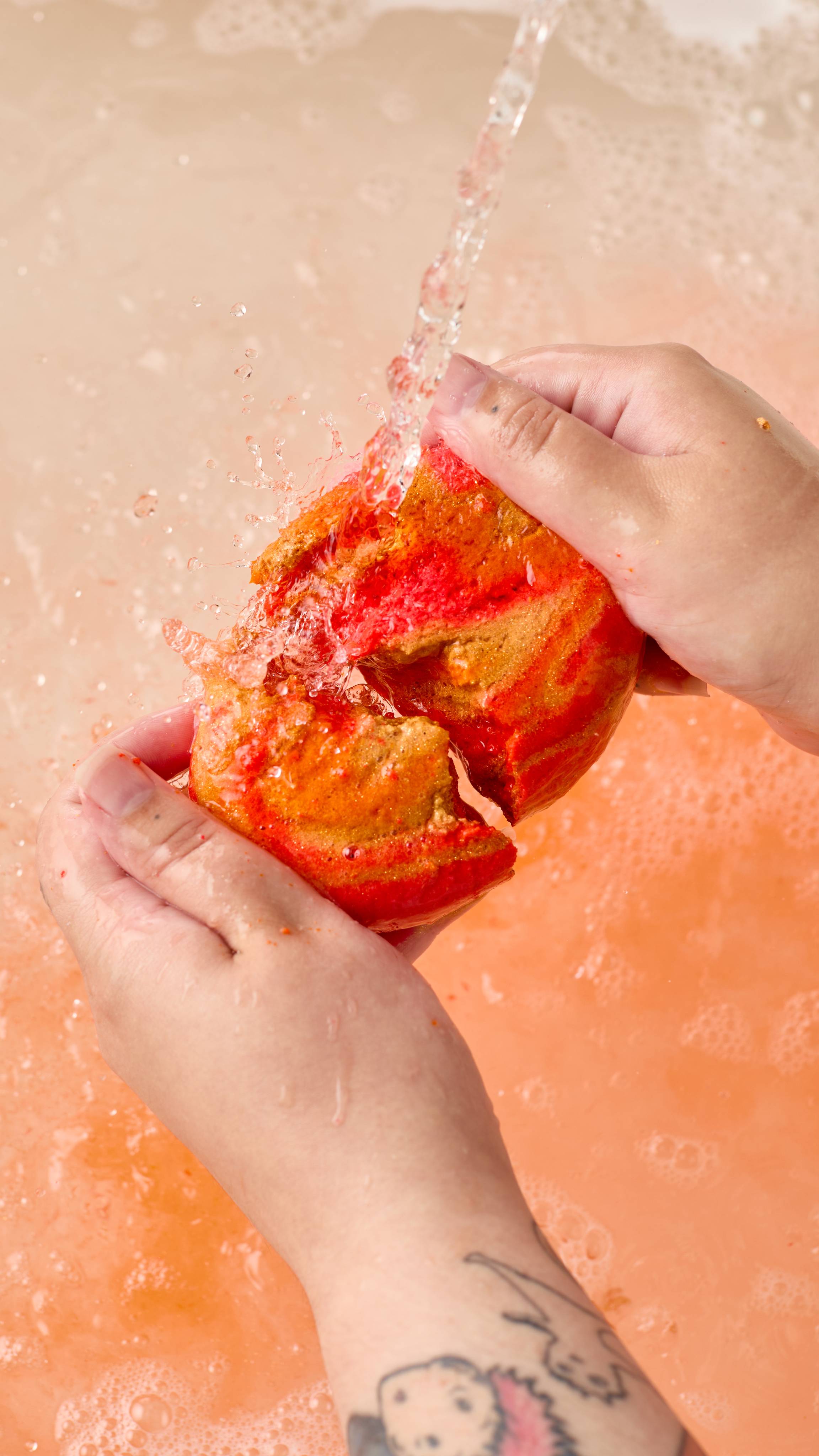 The image shows two light-toned hands vigorously breaking a vibrant gold and orange 'Pumpkin Spice' bubble bar under a strong stream of water. Abundant orange foam and splashes explode across the bath.