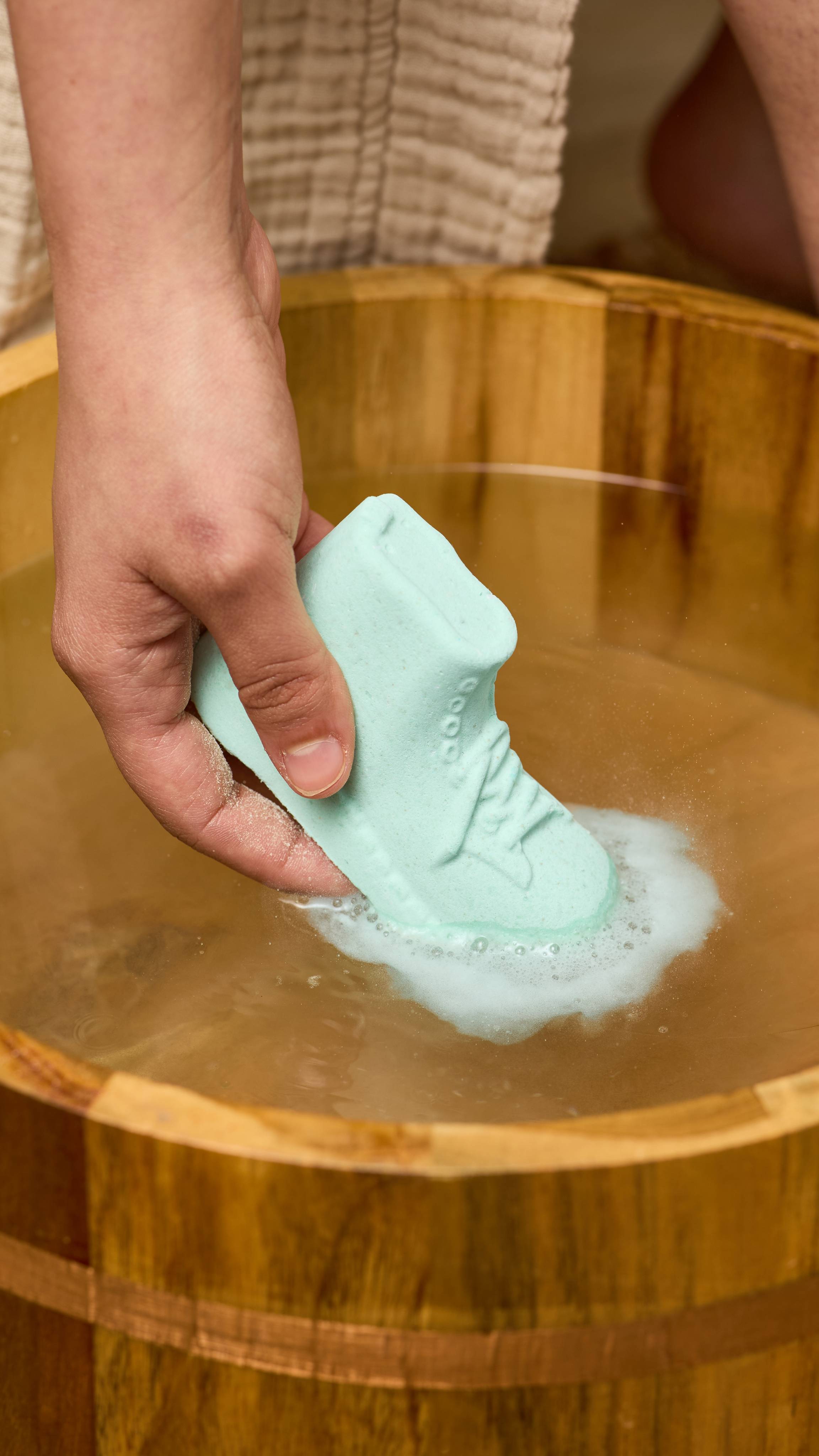 The image shows a light-toned hand holding the shoe-shaped, mint-green Put The Boot In foot soak product. White foam gathers around the product as it fizzes in a wooden tub filled with light-coloured water.