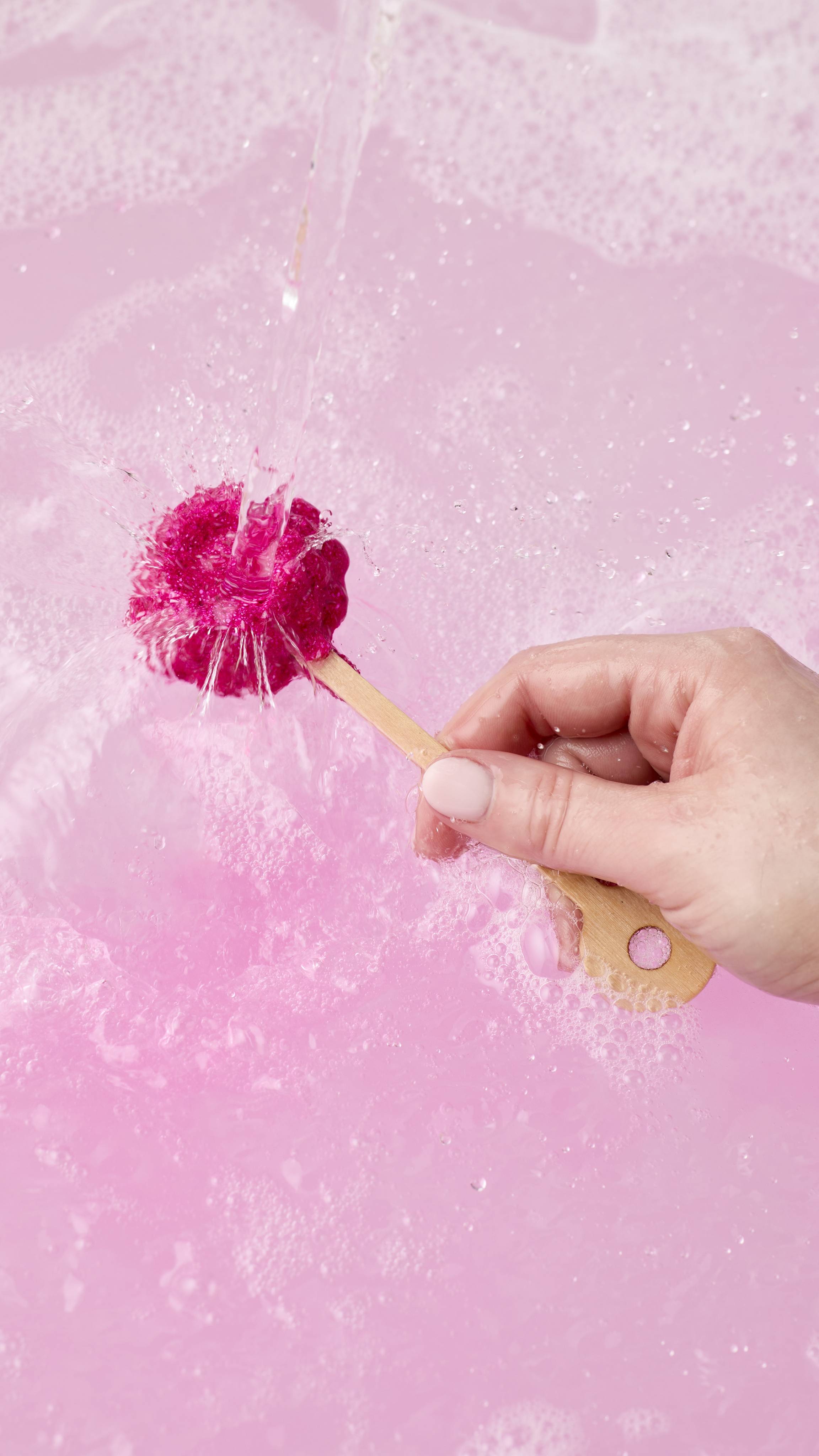 A hand holds a Raspberry mini bath wand under a stream of running water. The water splashes off the shimmering magenta base, creating vibrant pink swirls as it dissolves into the pale pink, foamy bath water below.