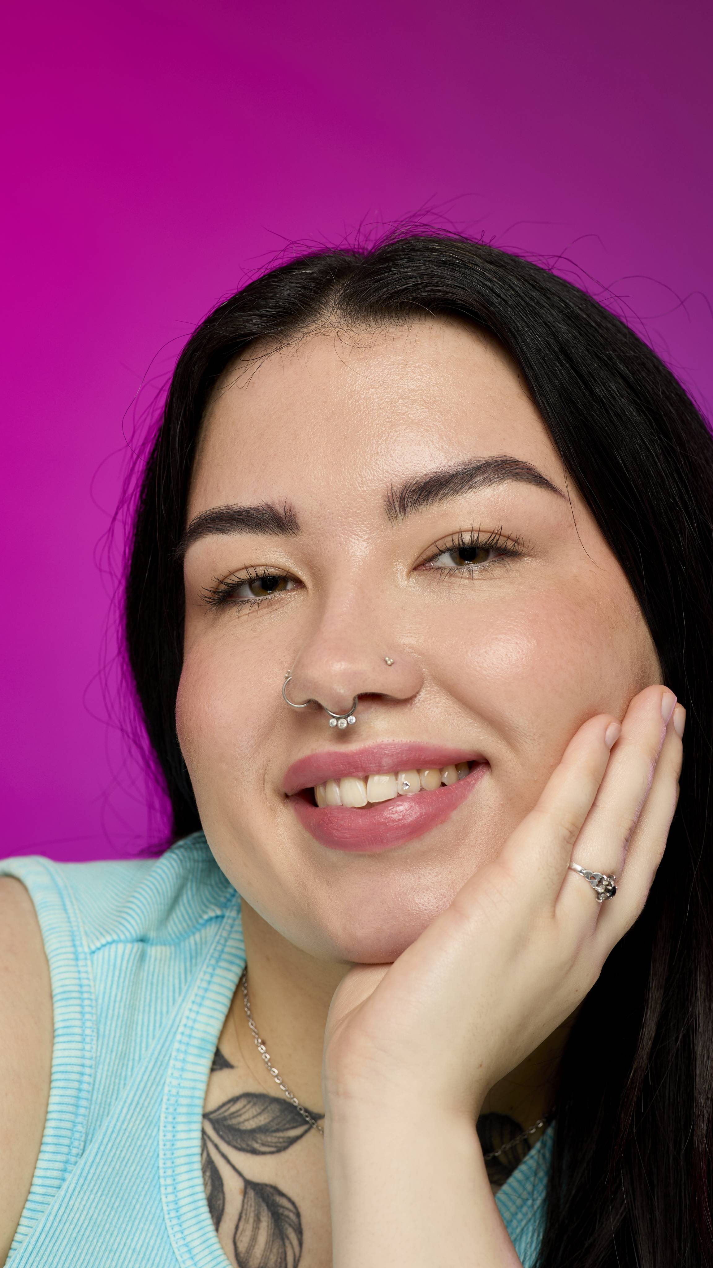 A smiling model with long, dark hair, wearing a light blue ribbed top, is shown from the chest up. They have their chin resting on their hand, and they are smiling to show off the velvety, subtle, rusted-red Roman Candle lipstick.