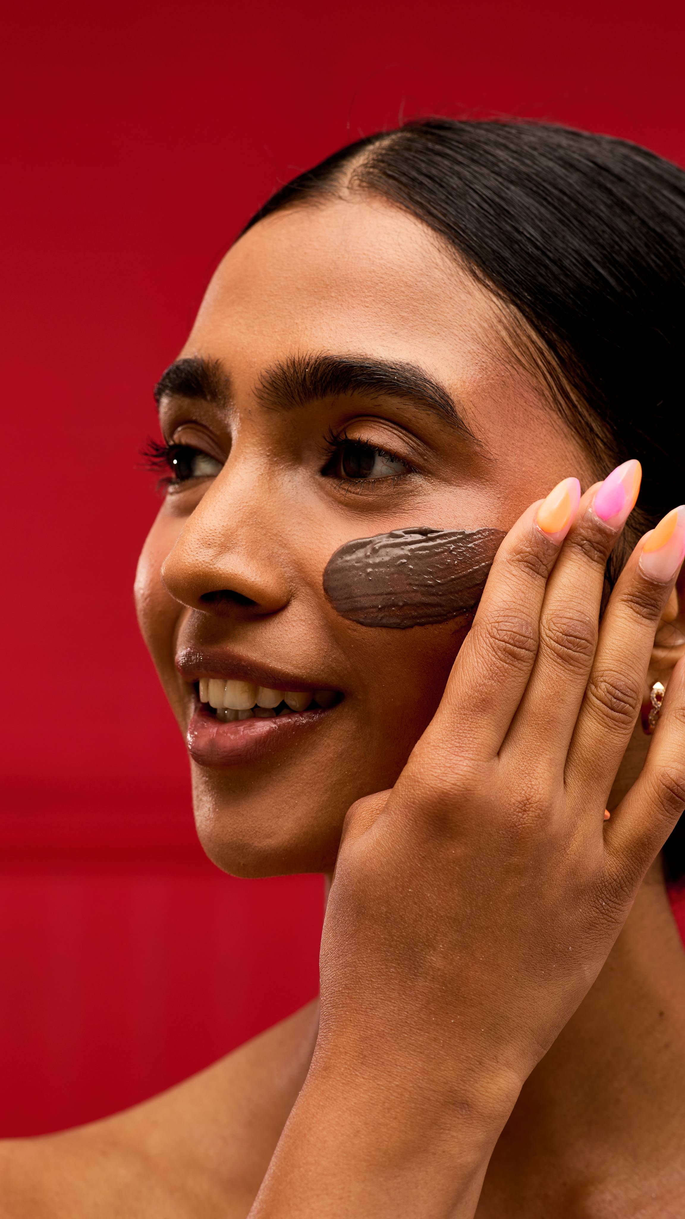 A person applies a dark brown, textured LUSH Rudolph jelly mask to their cheek. Their hand has a colourful ombré manicure, and the background is a vibrant red.