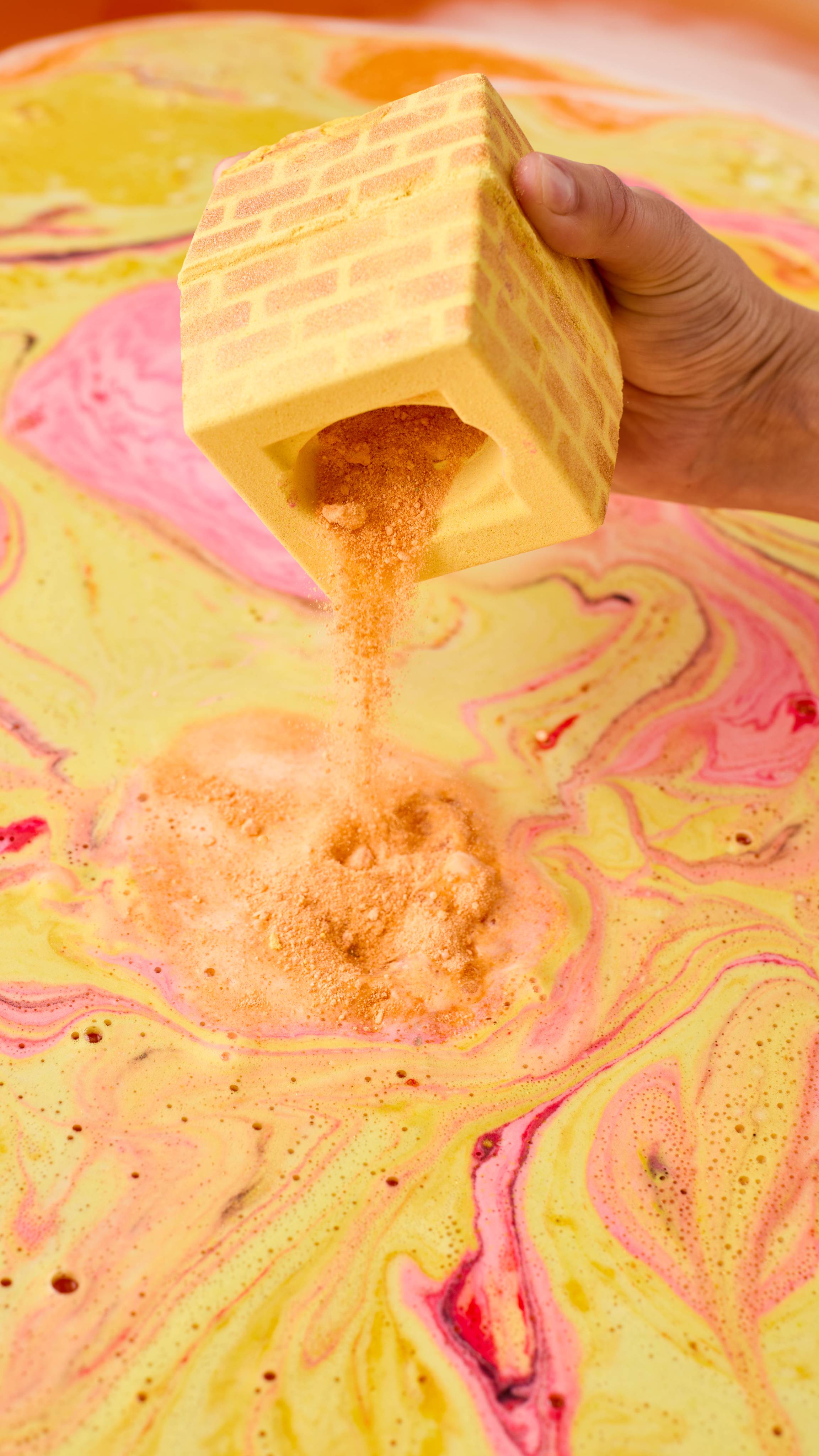 The image shows a hand holding the yellow, brick-patterned Stuck In The Chimney bath bomb. The legs ection has been removed and golden powder falls from the centre of the product into swirling bath water marbled with pink and yellow foam.