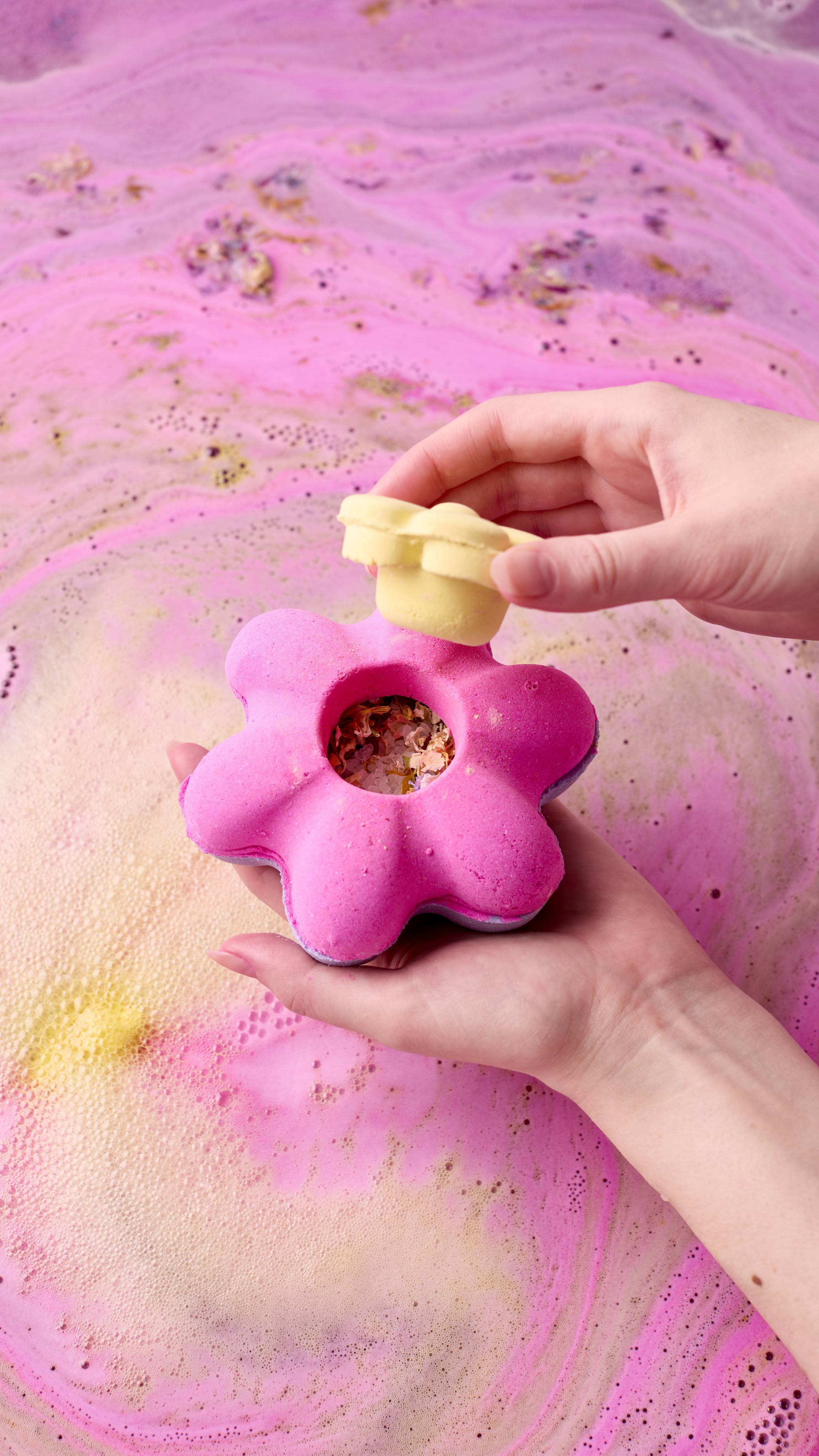 Hands lift the yellow flower-shaped lid from the magenta Secret Garden Bombshell bath bomb, revealing a hollow center filled with dried flower petals. The bath bomb is held above pink and yellow swirling bathwater.