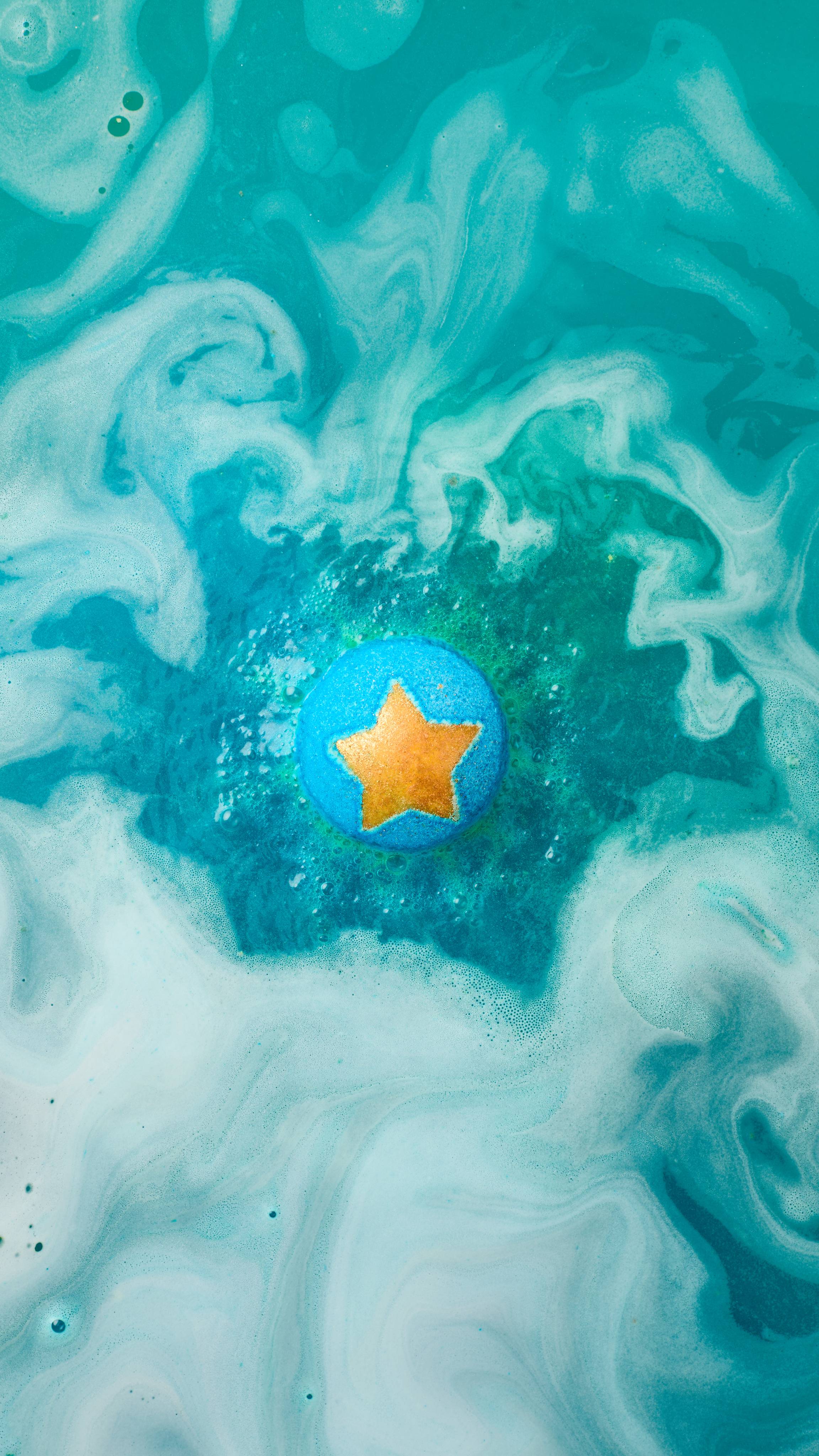 The image shows a blue, star-topped 'Shoot For The Stars' bath bomb fizzing in the water. Vibrant hues of dark blue and turquoise swirl around the product, mixing with abundant foam and bubbles.