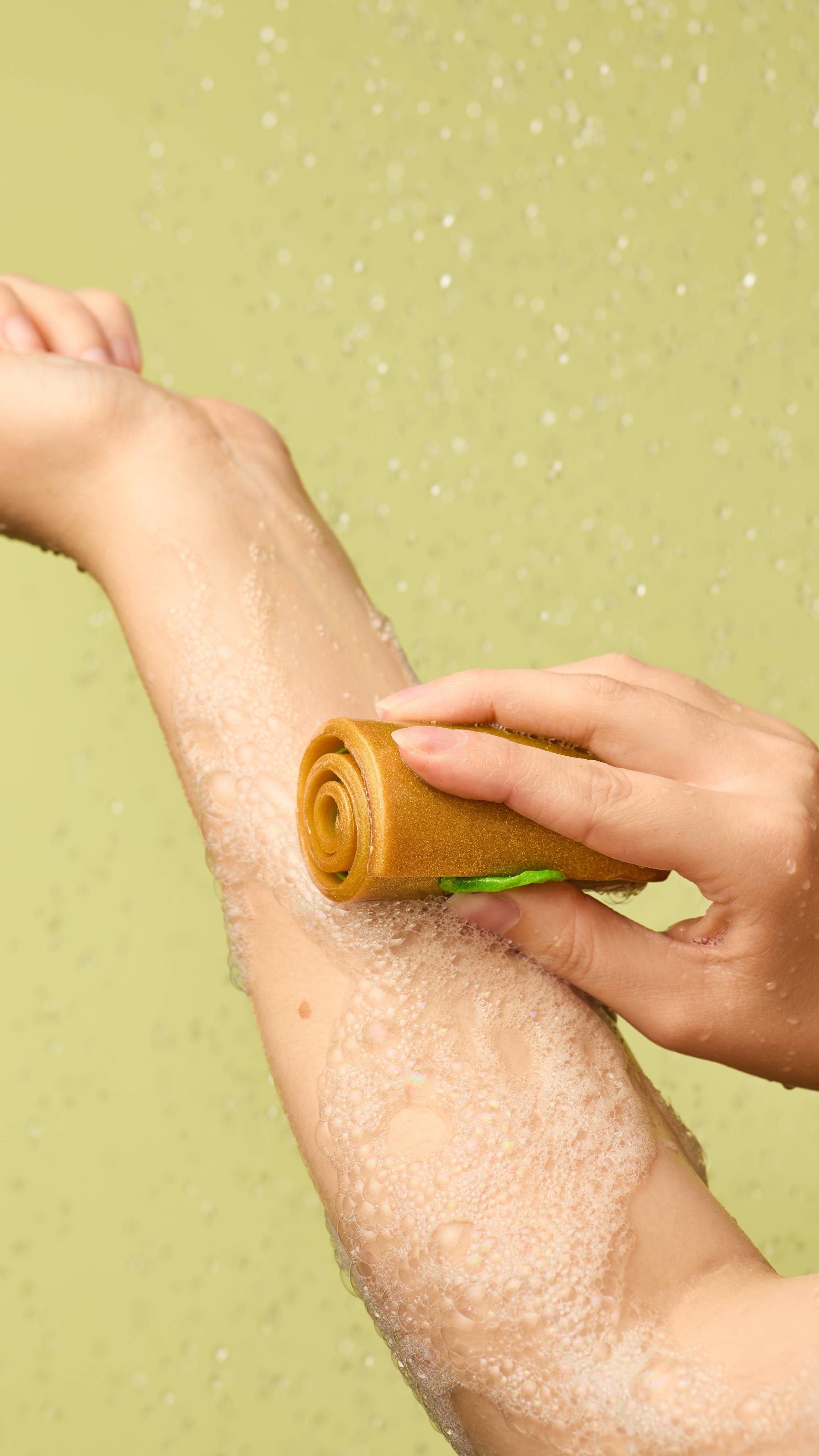 The image shows a hand rubbing a cylindrical, caramel-coloured Showstopper soap with golden lustre and a green interior onto an arm. White, bubbly lather forms on the arm. Water droplets fall on a light green background.