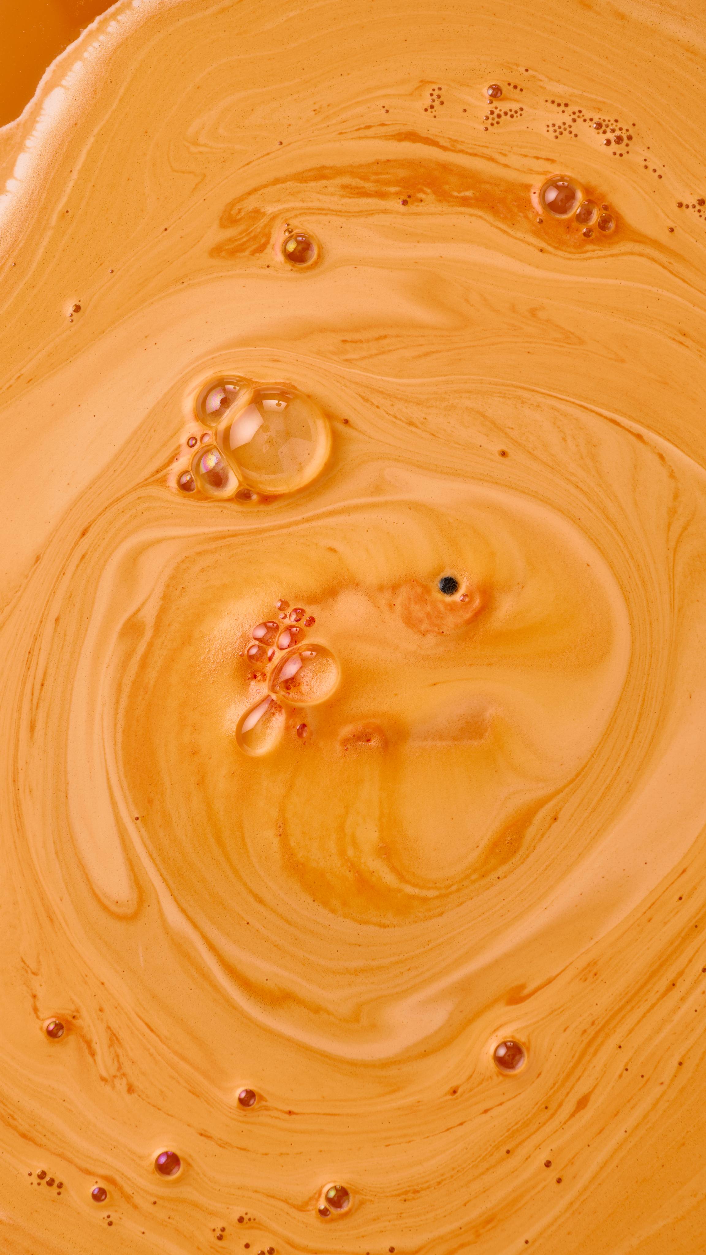 An aerial view of a bathtub filled with bright orange water and swirling, creamy foam, created by the "Shrimply The Best" bath bomb. Large, clear bubbles with iridescent reflections float on the surface of the velvety, orange foam.