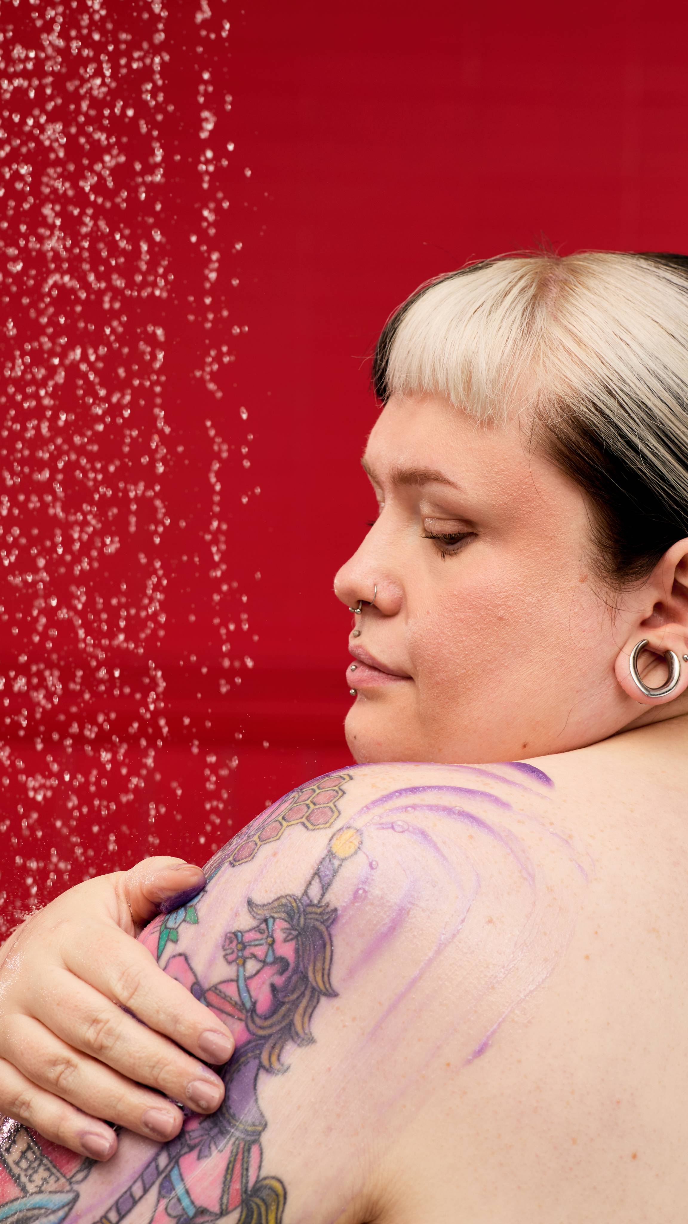 A person with two-toned hair and a tattoo on their shoulder applies a purple, shimmering LUSH Sleepy Bear shower gel. A fine mist of water droplets falls in the air, creating a sparkling effect against a vibrant red background.