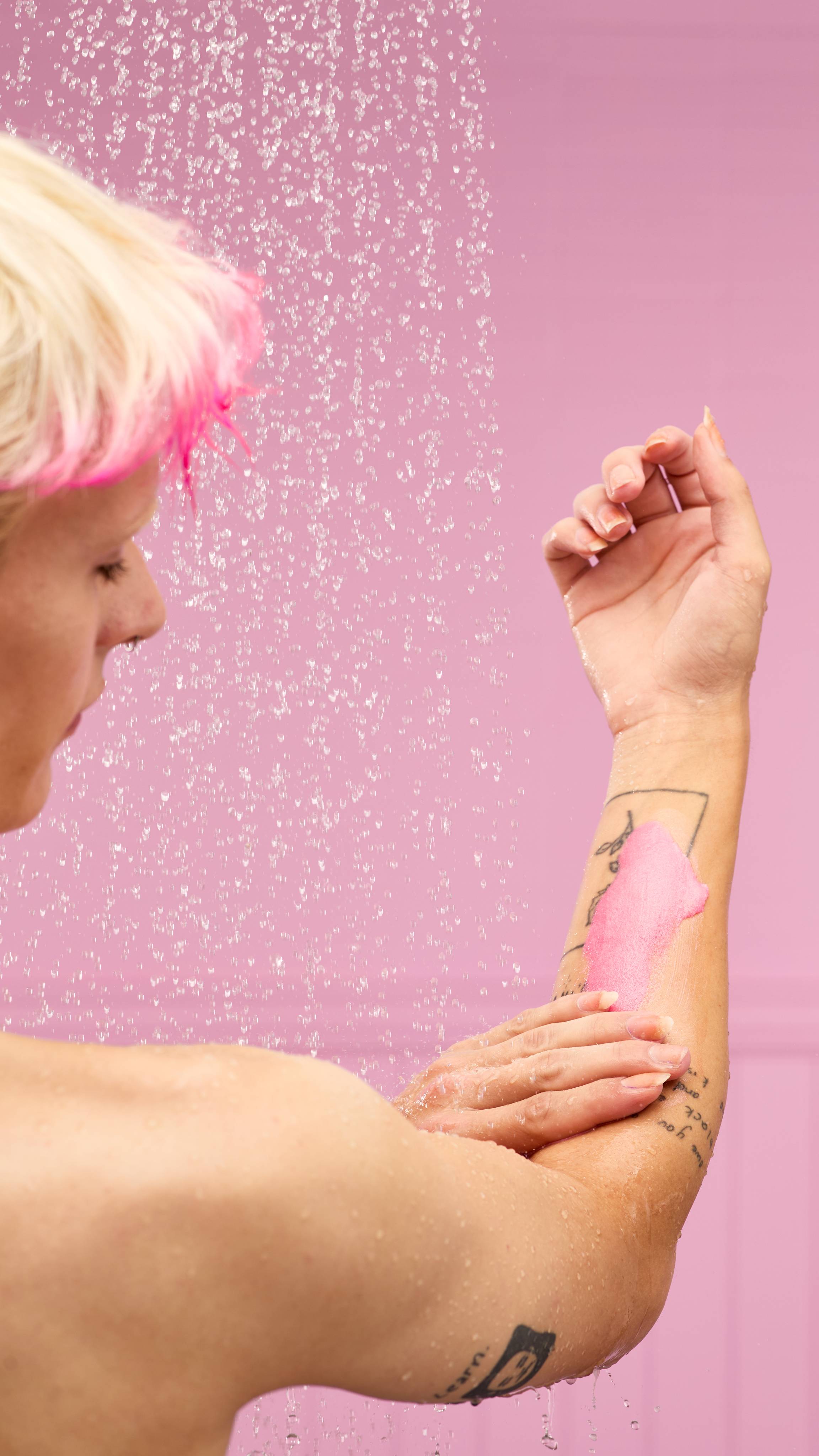 The image shows a solid, pale pink background with blurred, falling shower water. A light-toned arm reaches upwards as a hand lathers and exfoliates the forearm with the pink Snow Fairy body scrub.