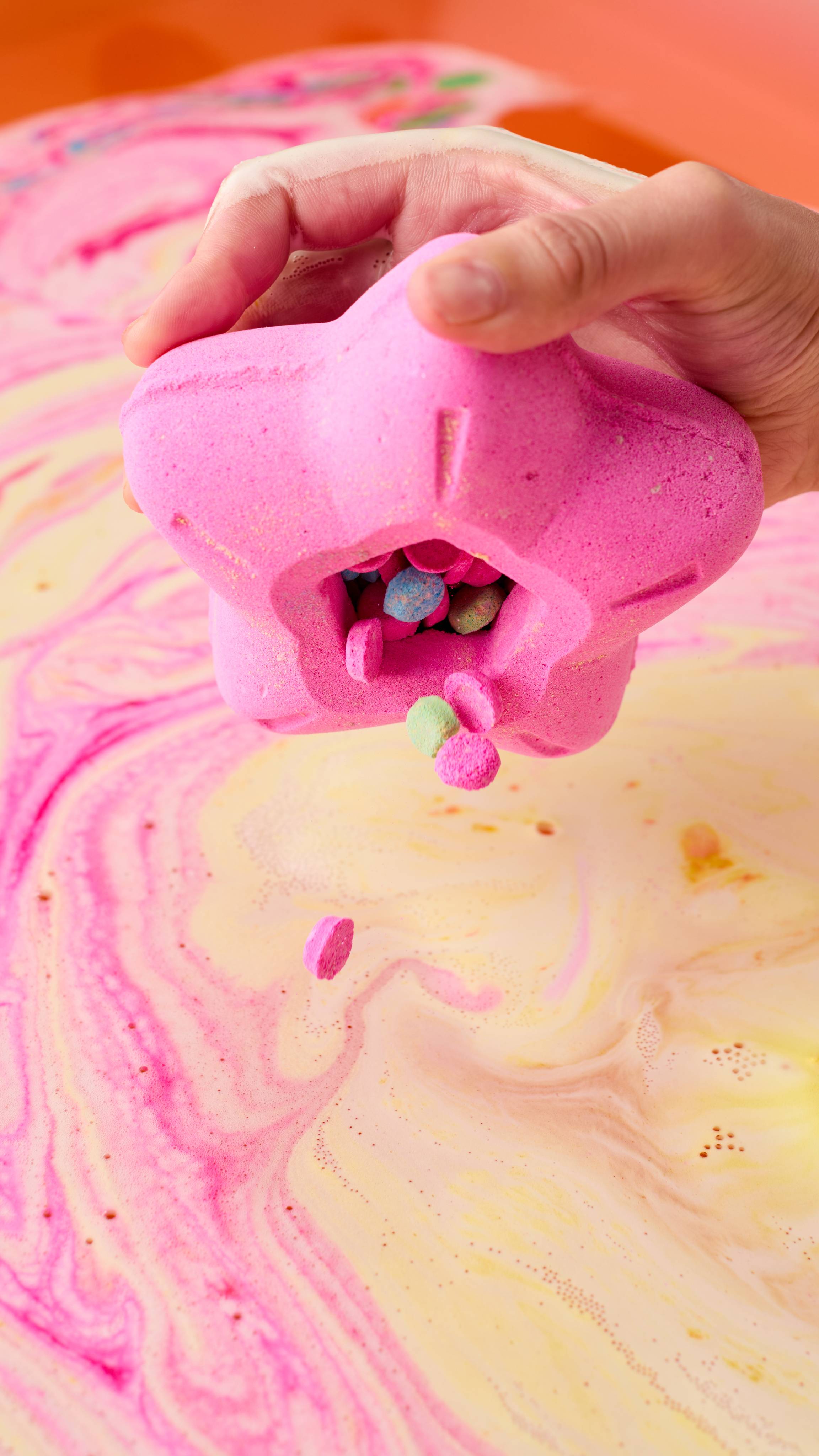 The image shows a hand holding the Snow Fairy Lights bath bomb. Small bath bomb confetti pieces of pink, blue, and green fall from the bomb into the swirling pink and yellow bath water, which is covered with light foam.
