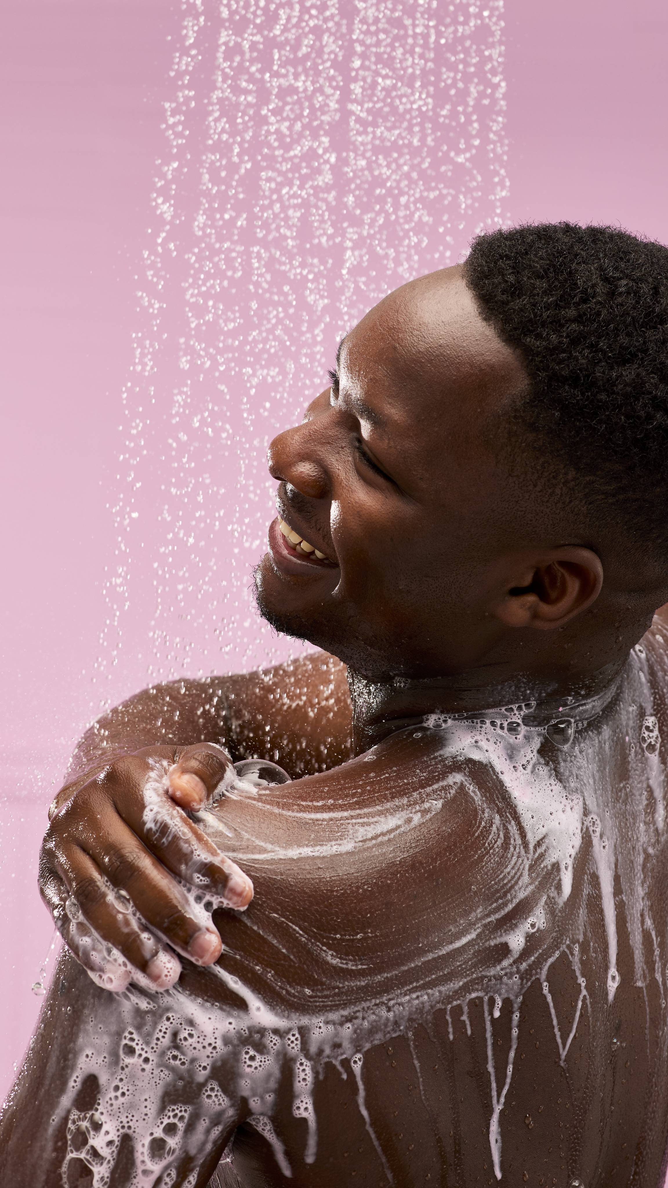A person with dark skin is shown from the chest up, in a shower against a pink background. They're smiling and looking away from the camera as they rub the Snow Fairy Milkshake shower gel into white lather over their shoulder.