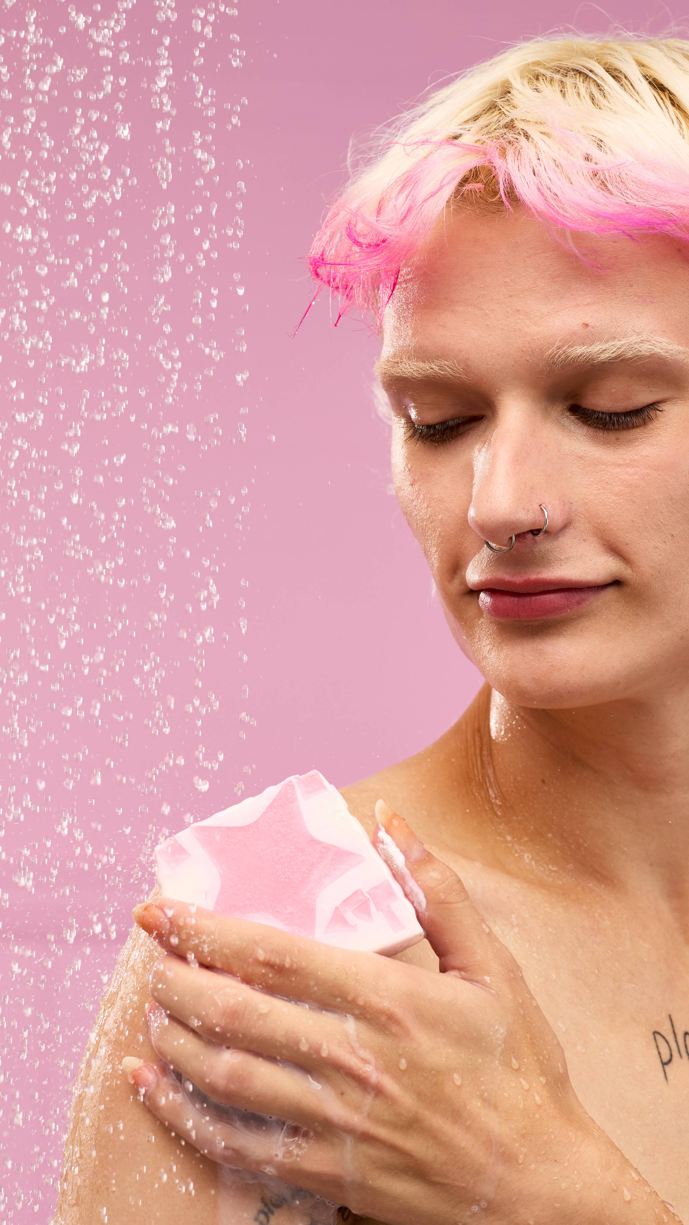 A person with bleached blonde hair and pink bangs is in a shower. Water streams down around them on a pastel-pink background. They hold the pink Snow Fairy soap slice and rub it on their left shoulder.