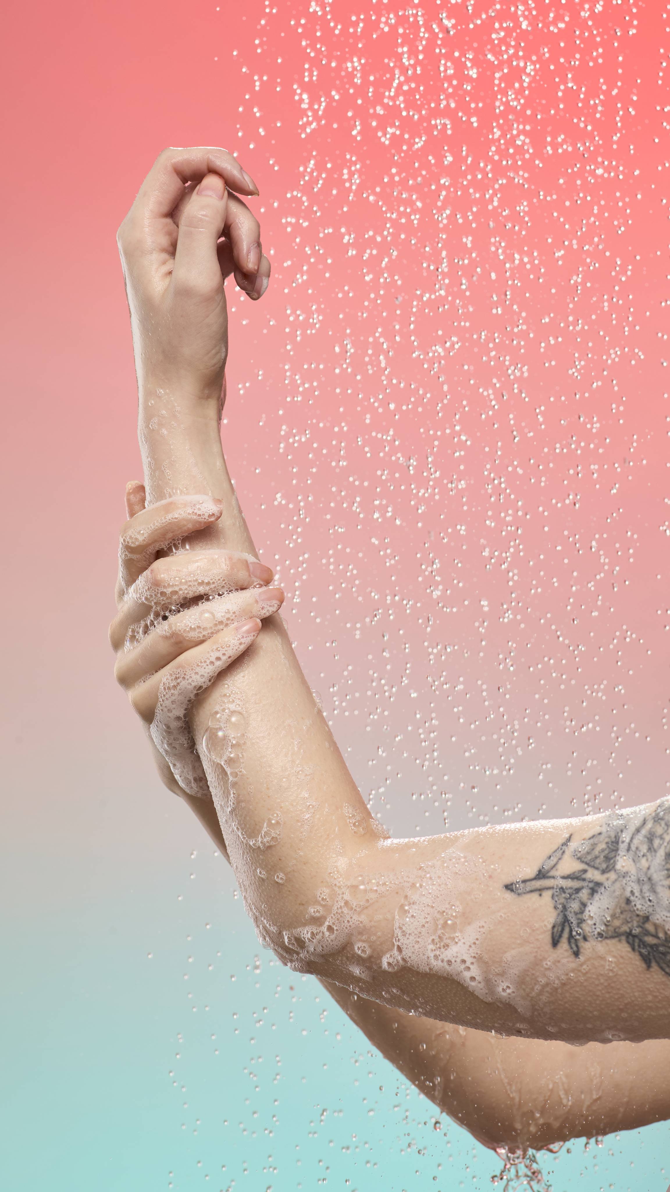 Hands lather a tattooed arm with white foam under a shower spray against a peach-to-blue gradient. Fine water droplets fall through the air around the soapy skin.