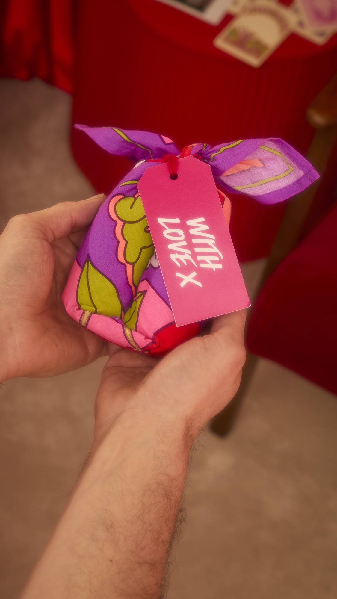 Hands hold the Squeeze Knot Wrap, displaying the purple fabric patterned with green leaves and yellow shapes. A bright pink tag with the message "WITH LOVE X" hangs from the bundle against a red background.