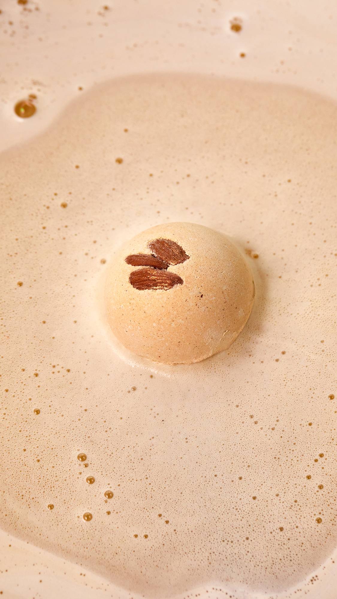 The image shows a close-up aerial view of the creamy Super Milk bath bomb as it starts to dissolve in the bath. The product is creating a thick blanket of creamy, ivory-coloured, velvety foam. 