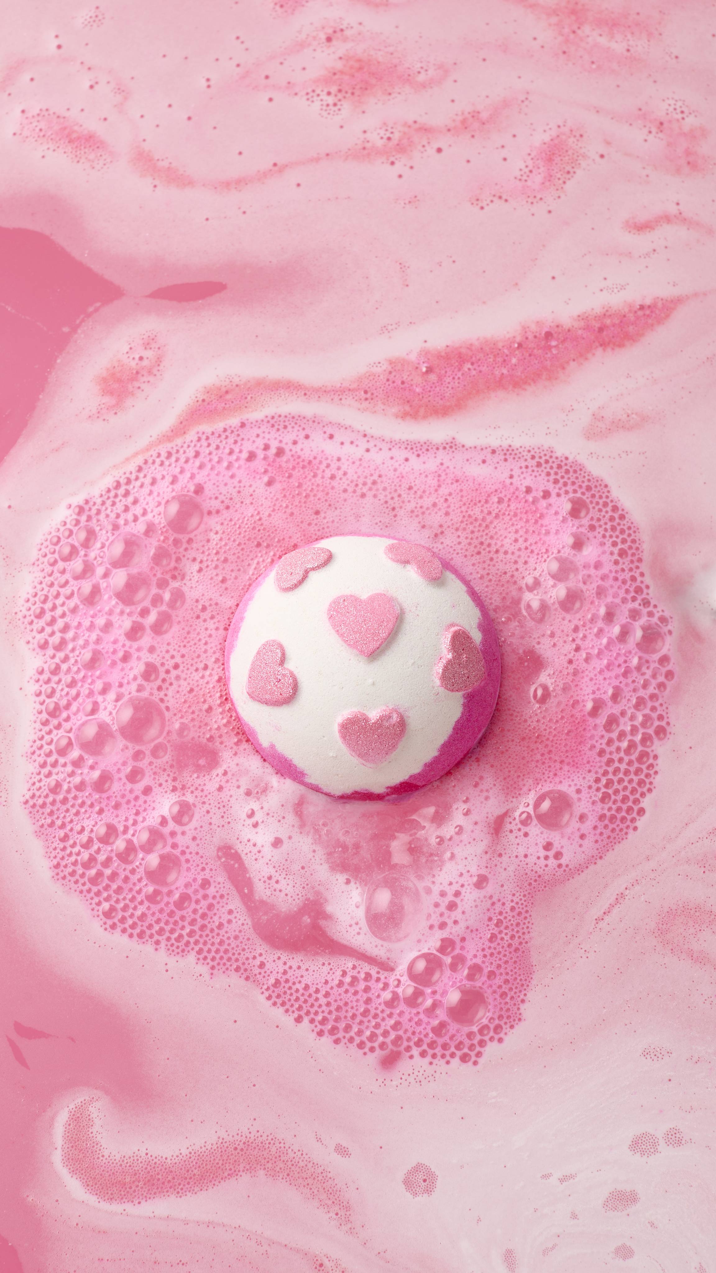 A white and pink Sweetheart bath bomb decorated with shimmery pink hearts dissolves in a bathtub, creating a halo of pink foam and bubbles against a background of swirling, pastel pink water.