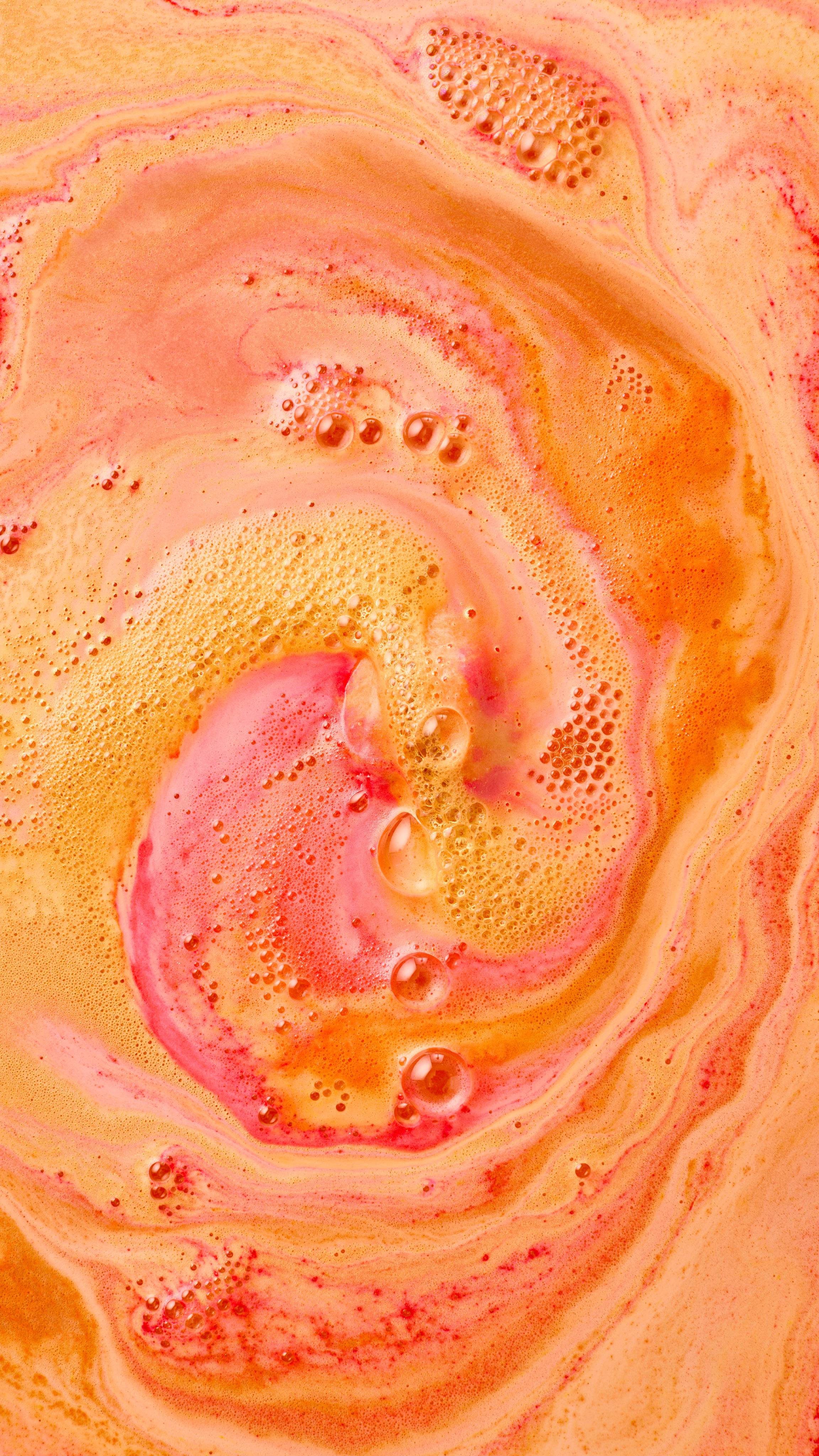 The image shows swirling bath art from 'The Alchemist' bath bomb, a vibrant blend of orange, yellow, and red-pink hues. Large, foamy swirls radiate from a central pink area, mixing with scattered bubbles across the water.