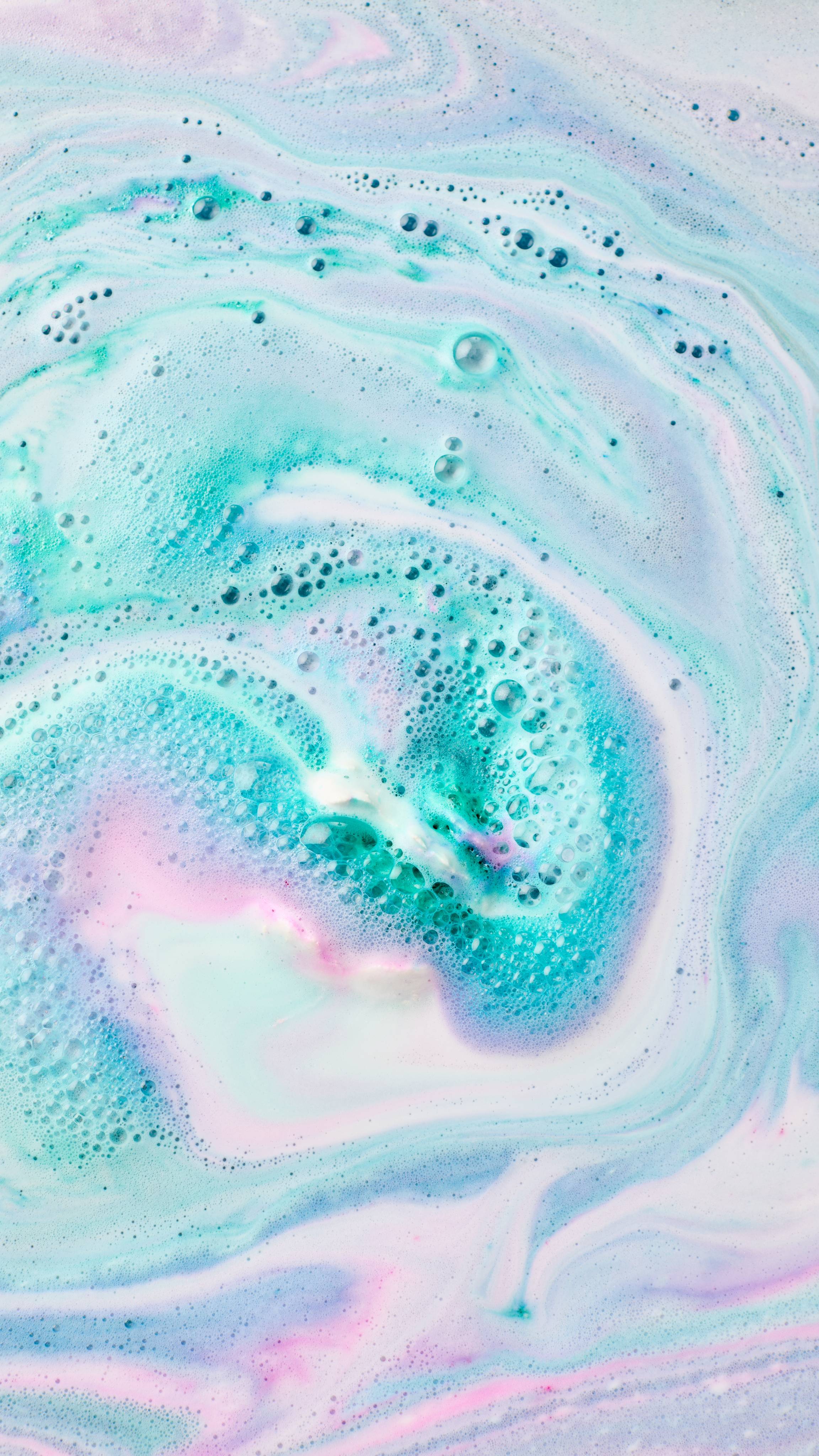 The image shows swirling bath art from 'The Girl In The Whirlwind' bath bomb, a vibrant blend of turquoise, pink, and white hues. Intricate marbled patterns mix with foam and bubbles.