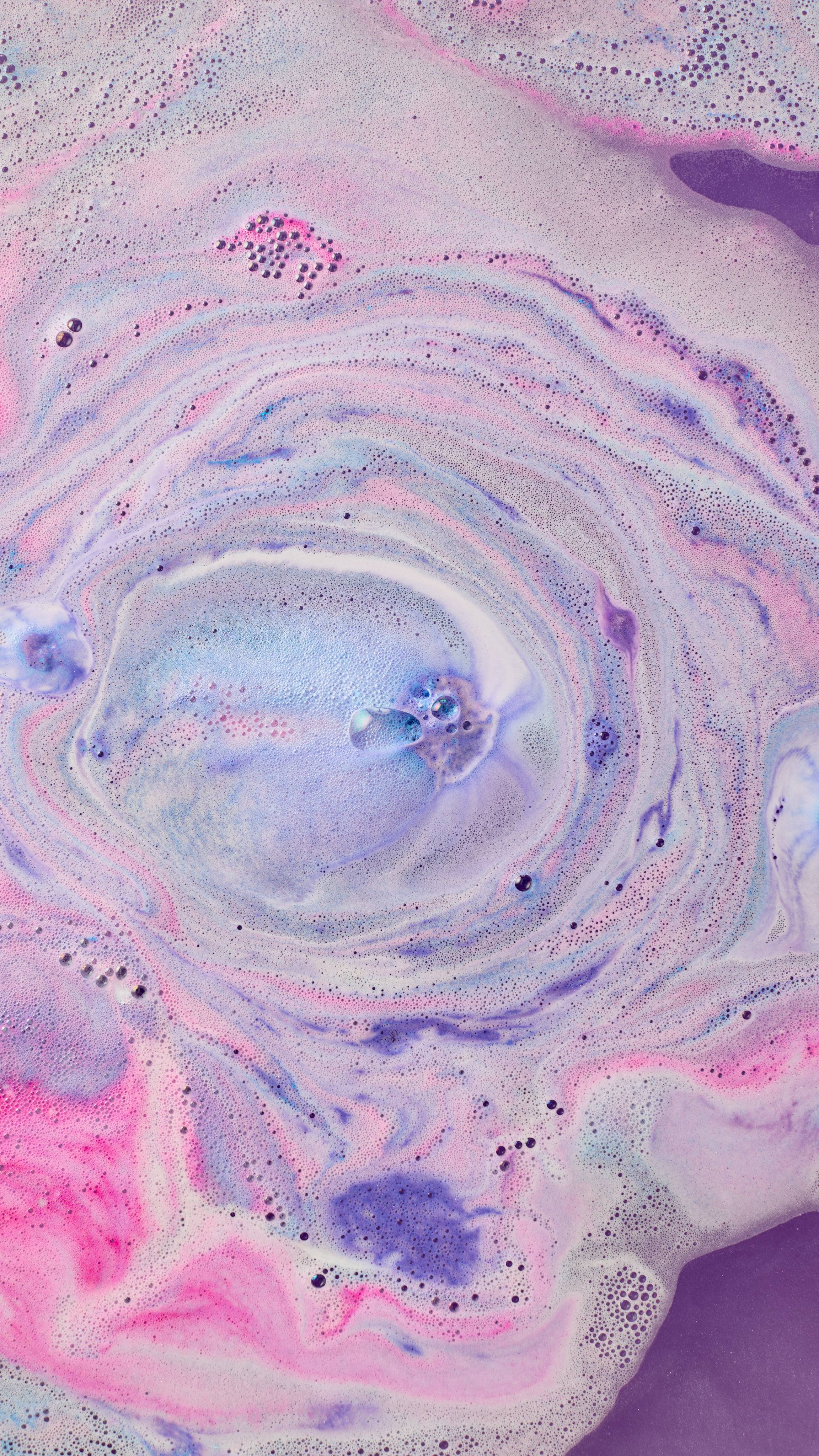 The image shows swirling bath art from 'The Magician' bath bomb, a dreamy blend of light purple, bright blue, and pink hues. Marbled patterns mix with darker spots and white foam across the water's surface.