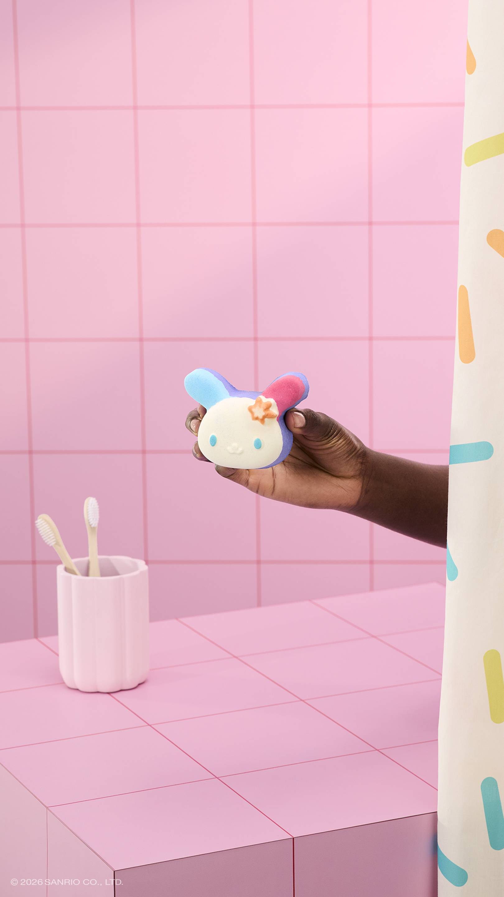 A hand holds a colourful bunny-shaped "U.SA.HA.NA" bath bomb in a pink tiled bathroom. A pink cup with two toothbrushes sits on the counter, and a sprinkle-patterned shower curtain is visible on the right. 